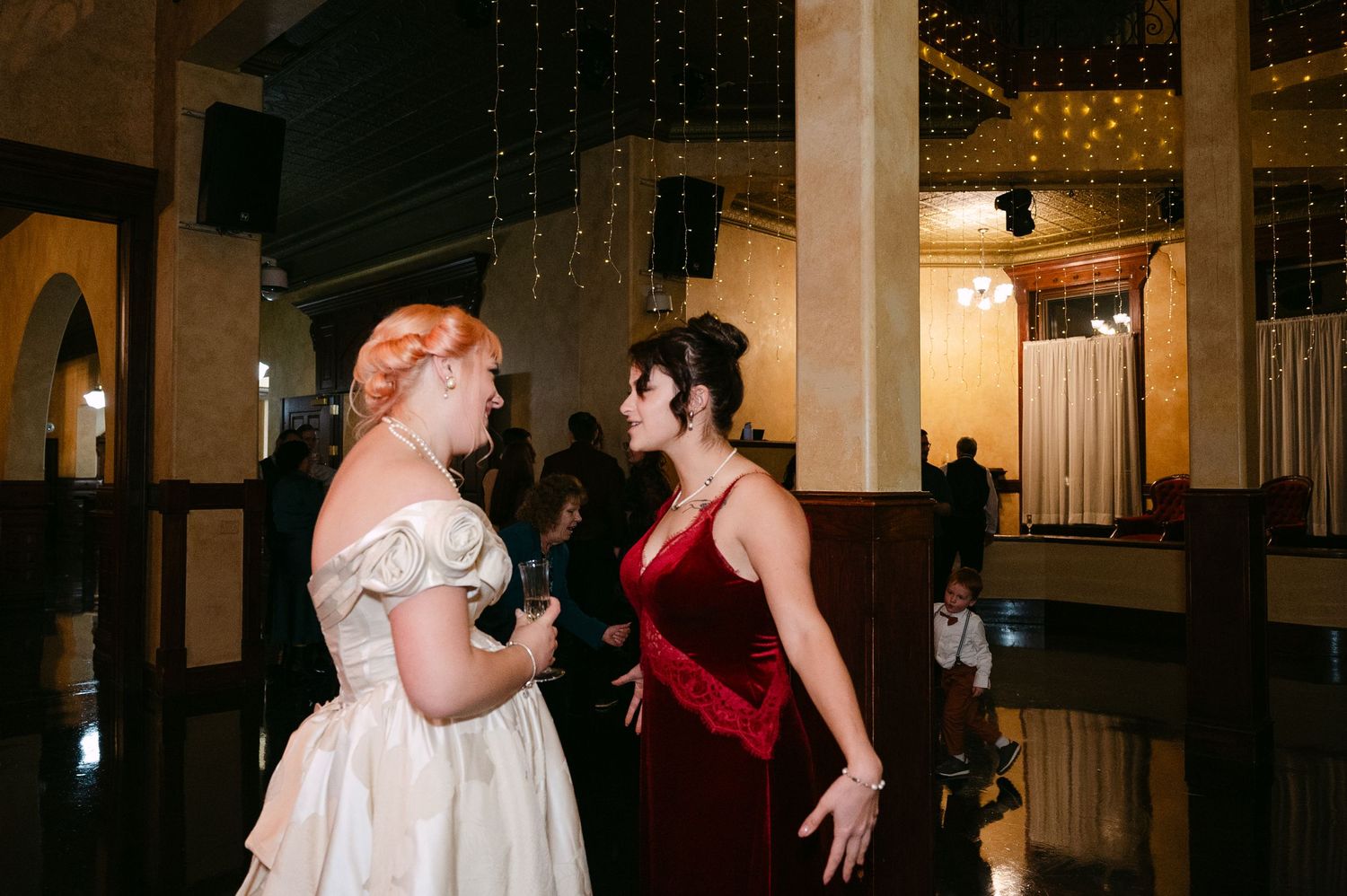 Central MN Wedding Photos | Bride in off-shoulder gown conversing with guest in red velvet dress.