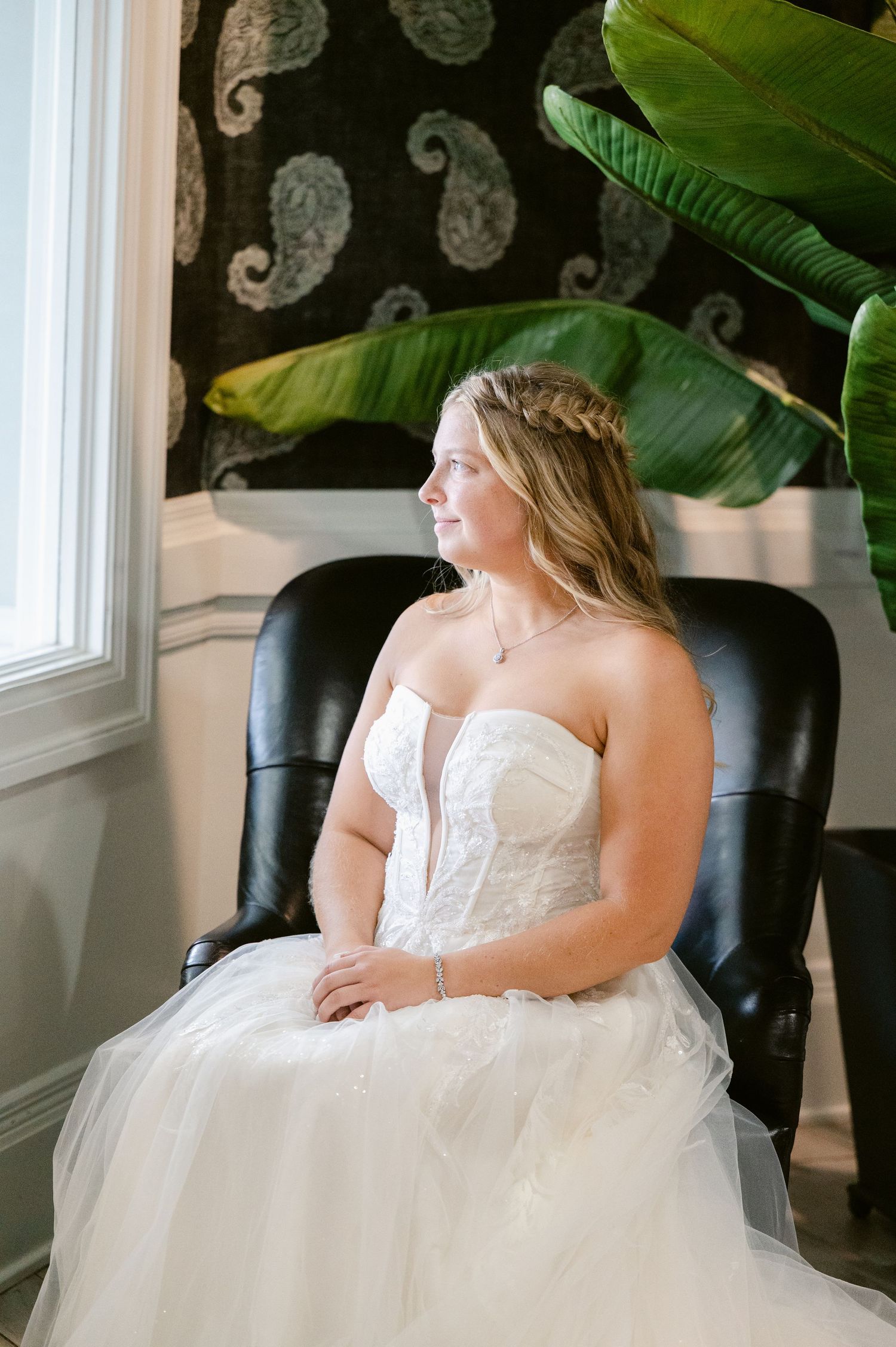 Central MN Wedding Photos | Bride in strapless lace tulle gown sits in black leather chair gazing out window with tropical plant.