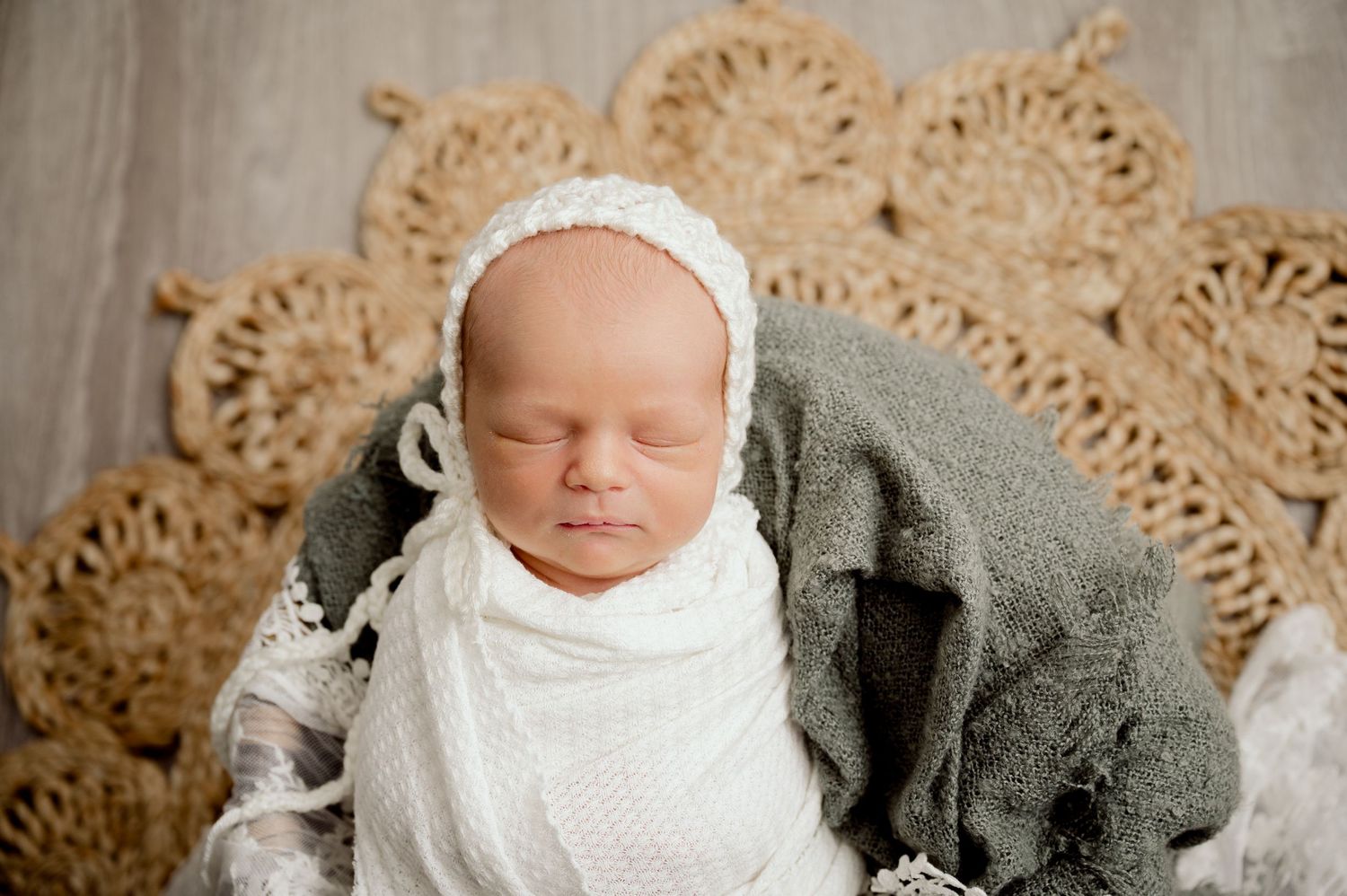 Sleeping newborn baby wrapped in white lace swaddle wearing knit bonnet on woven backdrop.