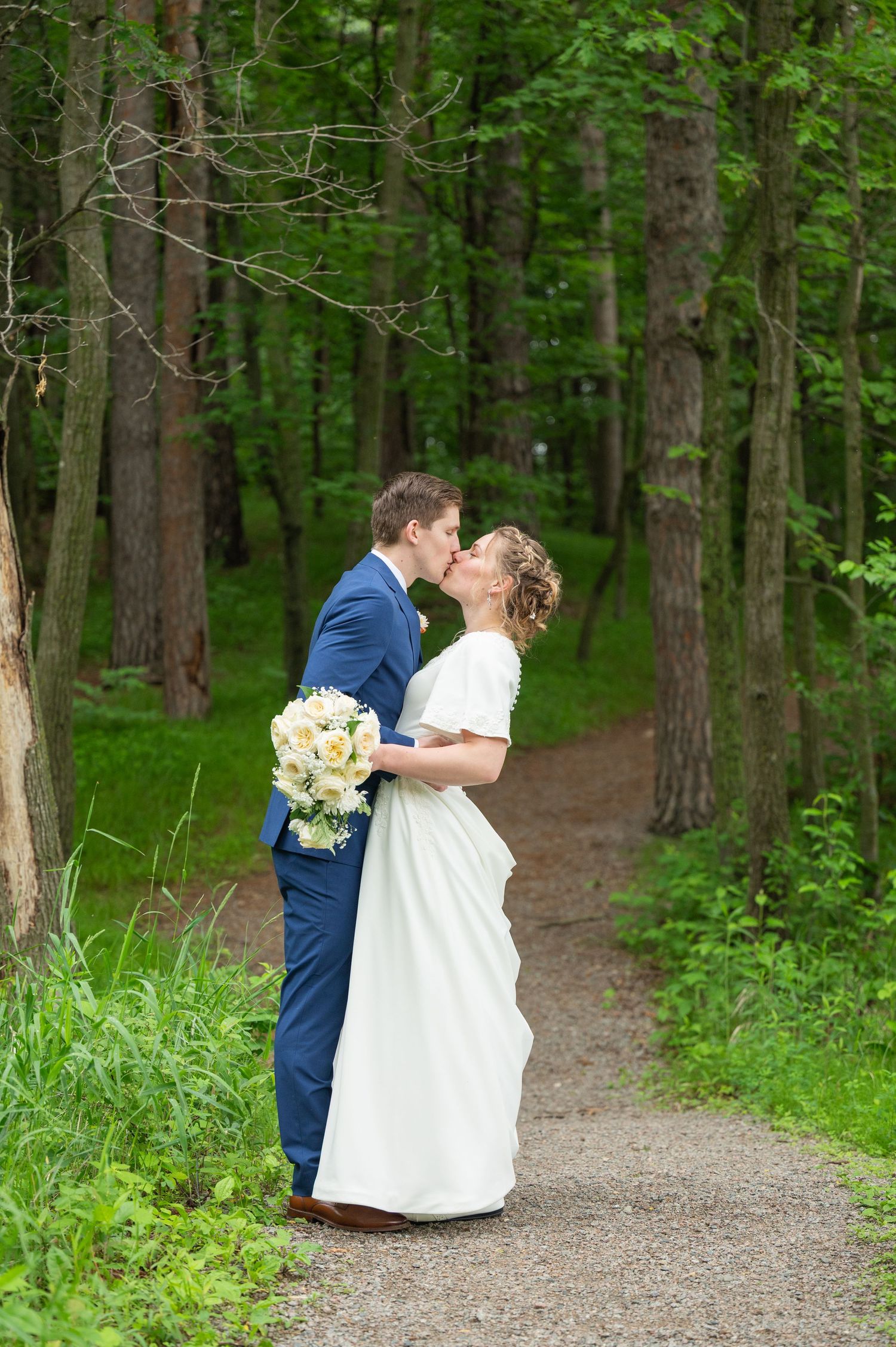 A romantic moment captured on a forest path surrounded by tall pine trees and greenery.