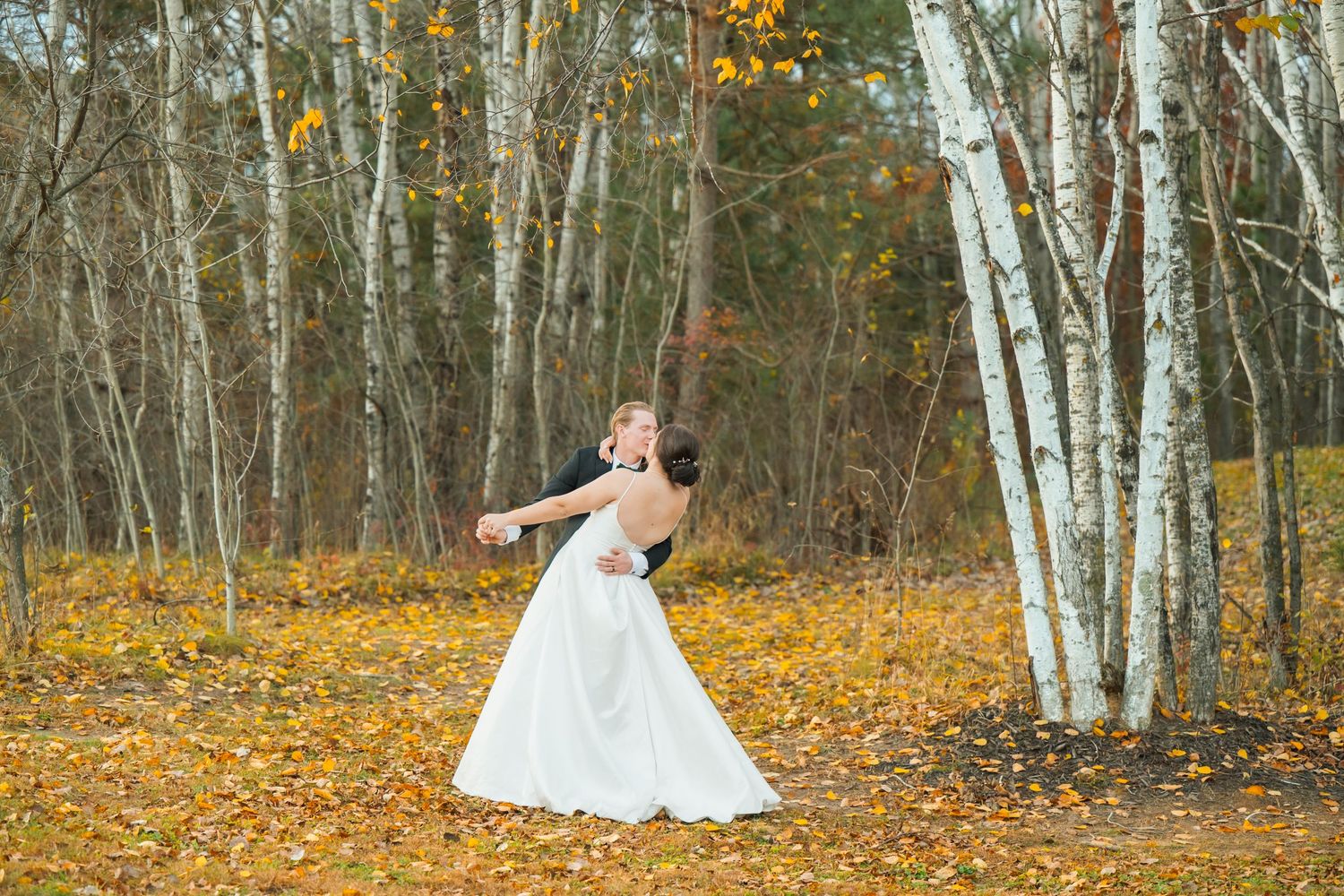 A bride in a white dress twirls joyfully among yellow autumn leaves and birch trees.