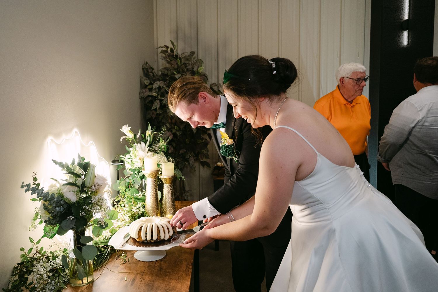 A couple in wedding attire cuts into their wedding cake together at a rustic wooden table decorated with white flowers.