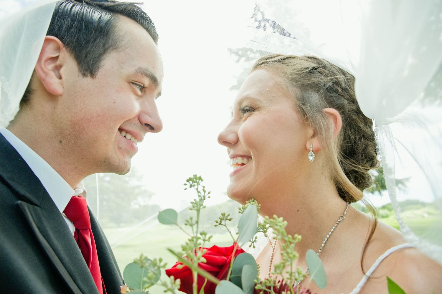 Smiling bride and groom share a romantic close-up moment under a veil with red rose bouquet outdoors.