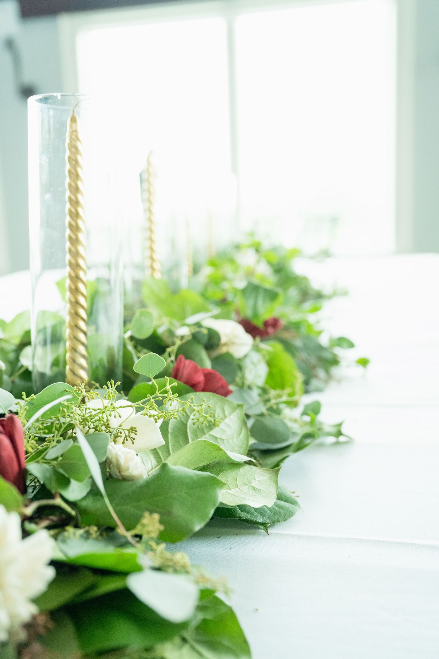 Elegant wedding table centerpiece with lush greenery, red berries, white flowers, and gold spiral candles on a white tablecloth.