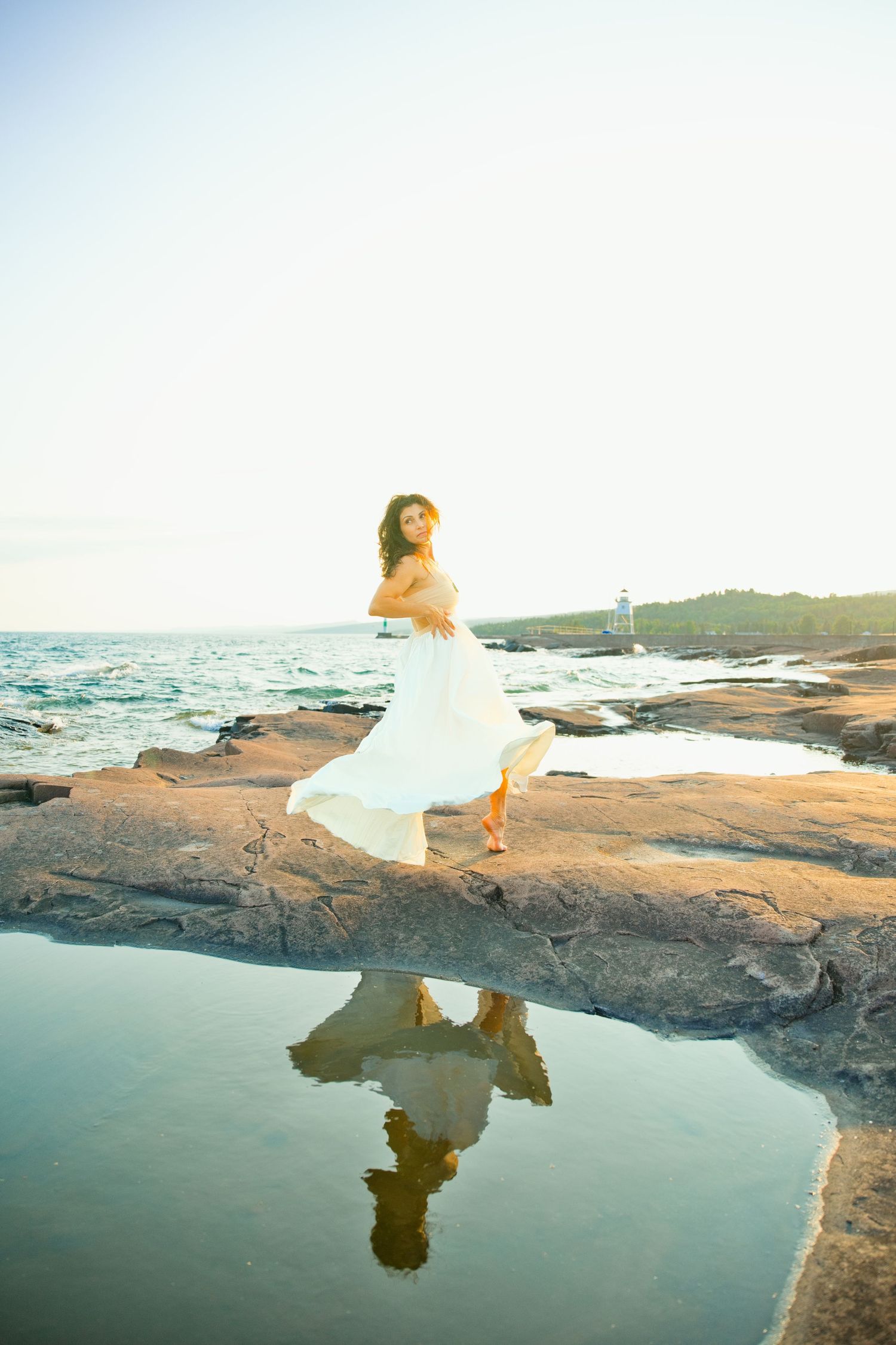 Woman in flowing white dress twirls on rocky shore with her reflection visible in a tide pool.