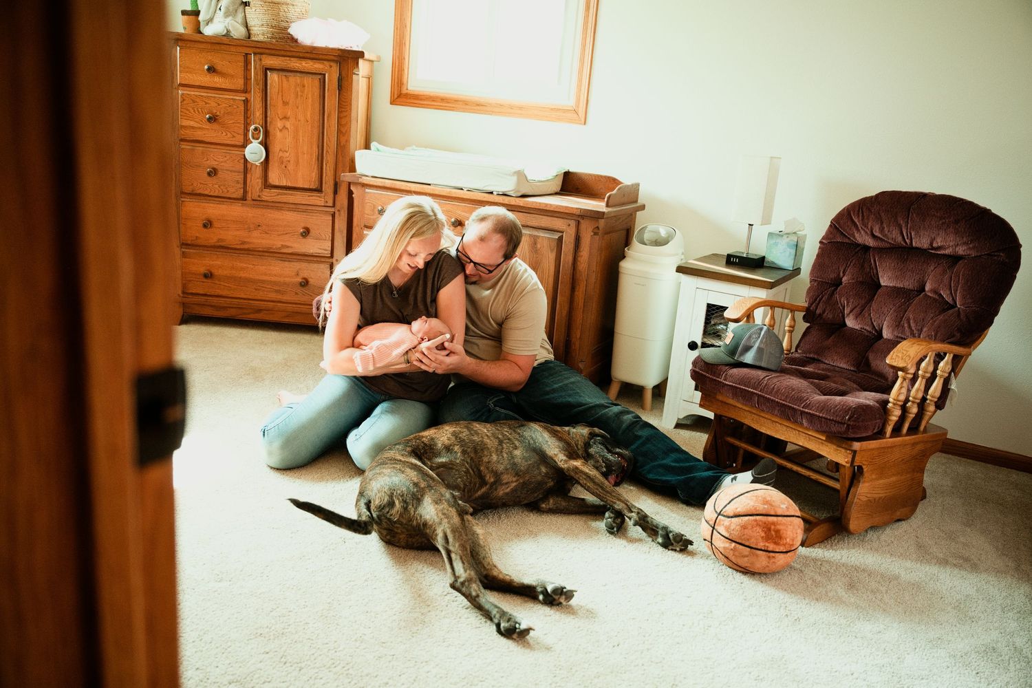 Couple sitting on floor cuddling with large brindle dog lying beside them near a basketball in a cozy living room.