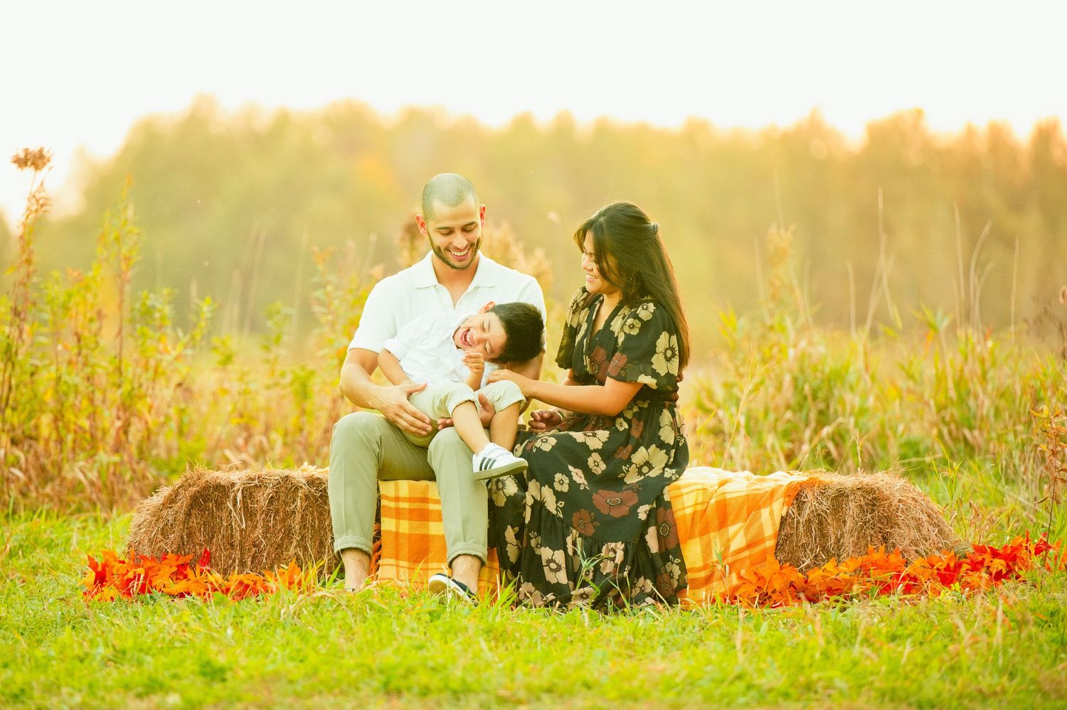 Family of three enjoying a warm autumn day outdoors on hay bales in a sunlit field with golden foliage.