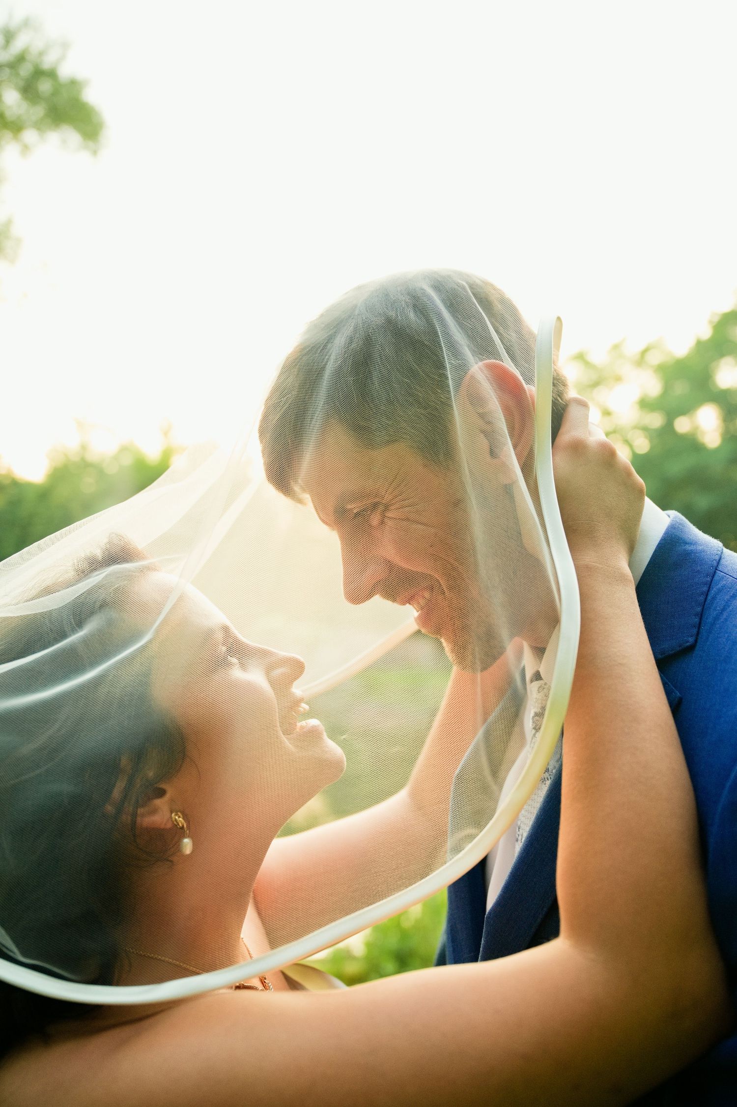 Bride and groom share romantic moment under wedding veil, smiling face to face outdoors in soft sunlight.