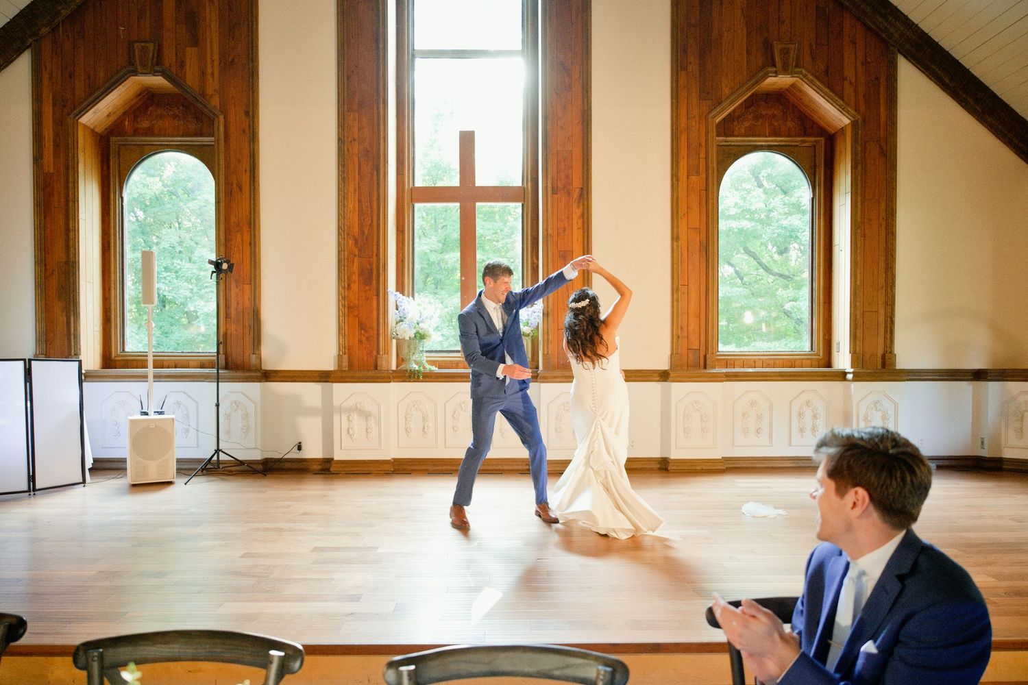 Bride and groom share first dance in elegant wood-paneled wedding venue while guest applauds in foreground.