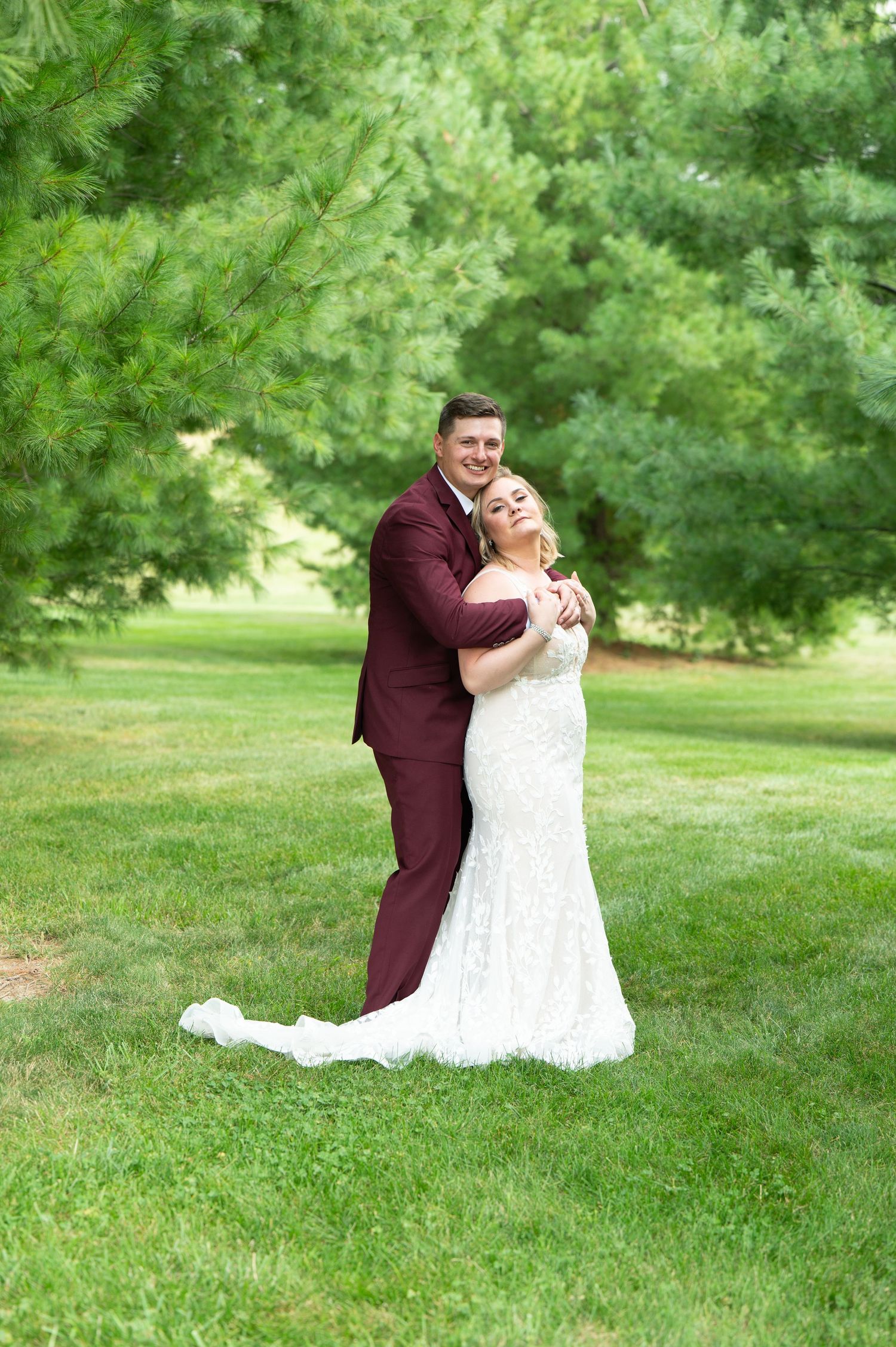 A newlywed couple embraces on a lush green lawn surrounded by trees at an outdoor wedding venue.