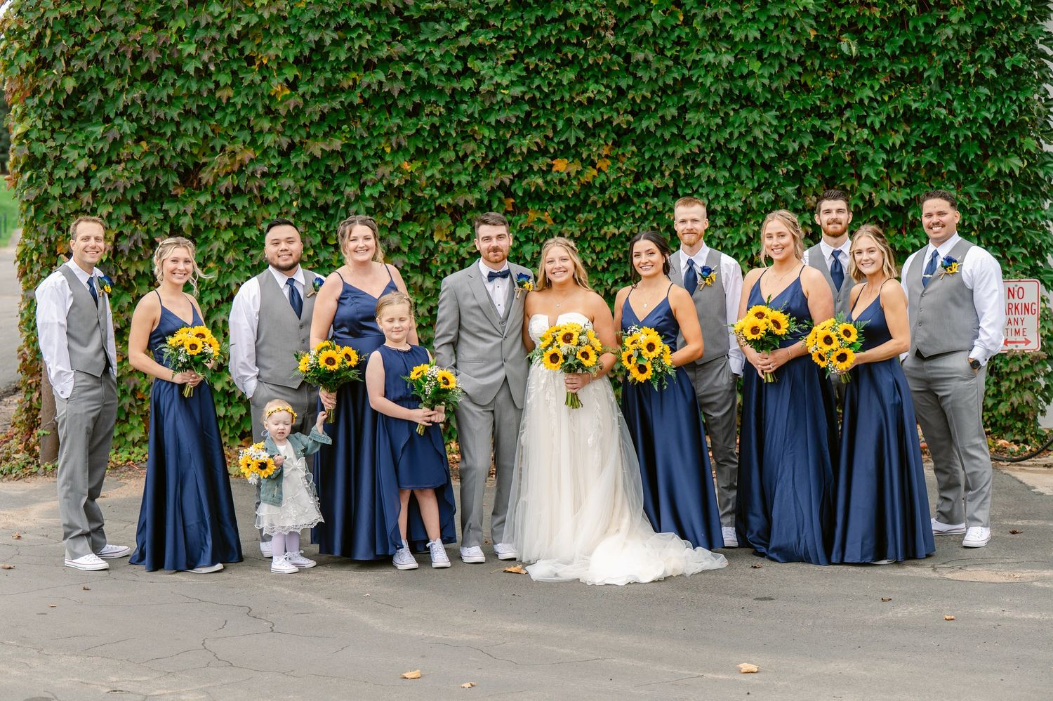 Central MN Wedding Photos | Wedding party poses in navy blue and gray attire with sunflower bouquets before ivy wall.