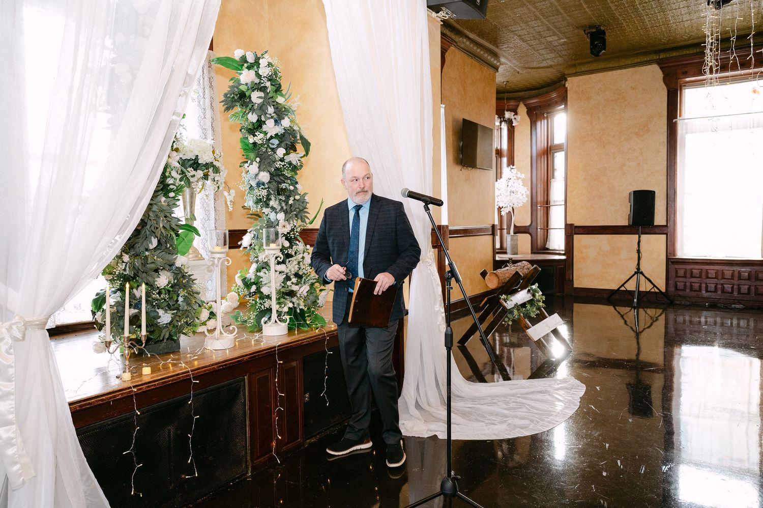 Central MN Wedding Photos | Elderly man speaking at a microphone podium decorated with white floral arrangements at a wedding.