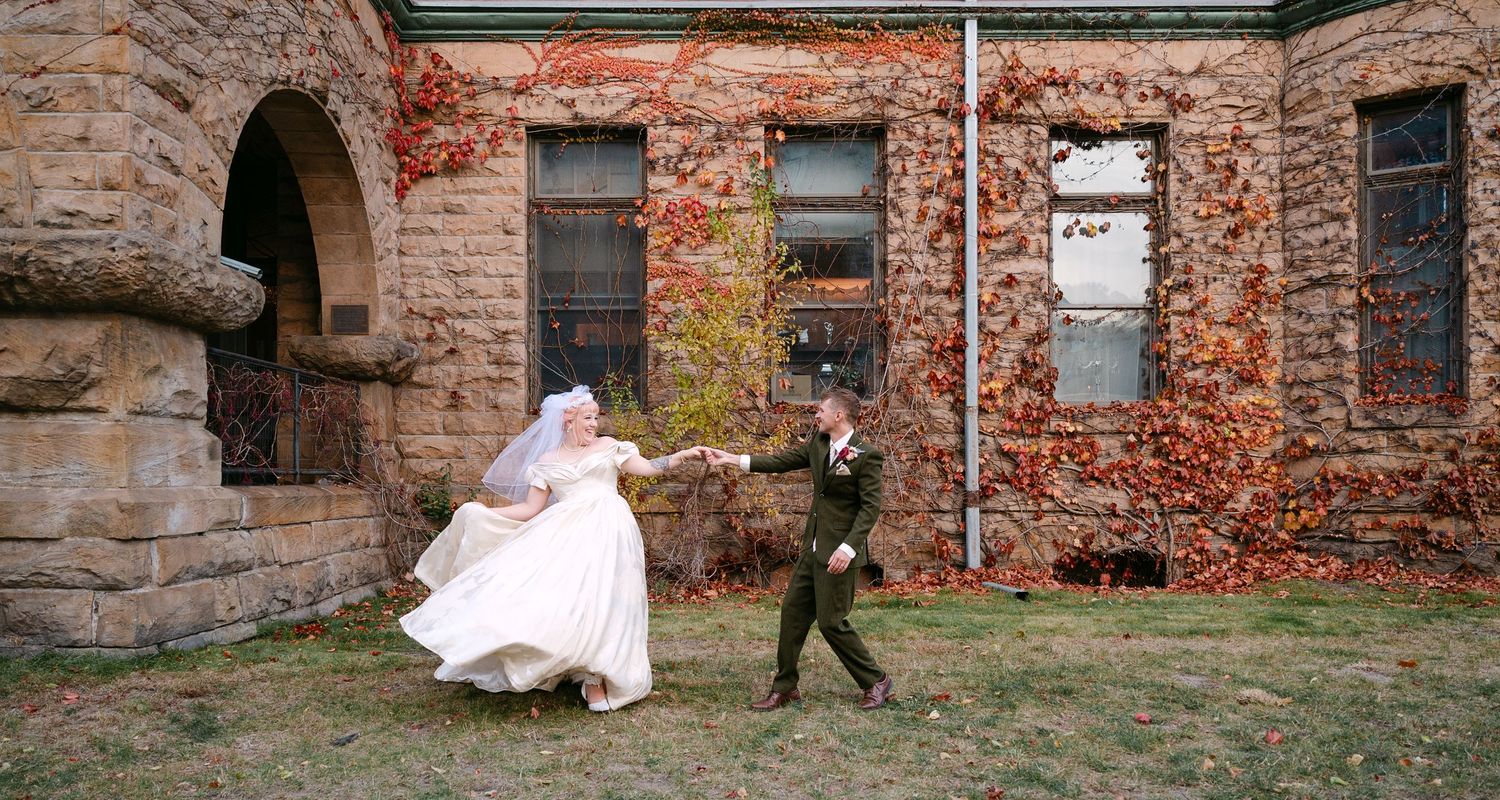 Central MN Wedding Photos | Bride and groom dancing playfully outside historic stone building with autumn red ivy walls.