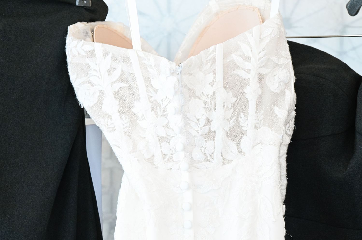 A delicate white lace wedding dress hangs on display in a bright room.