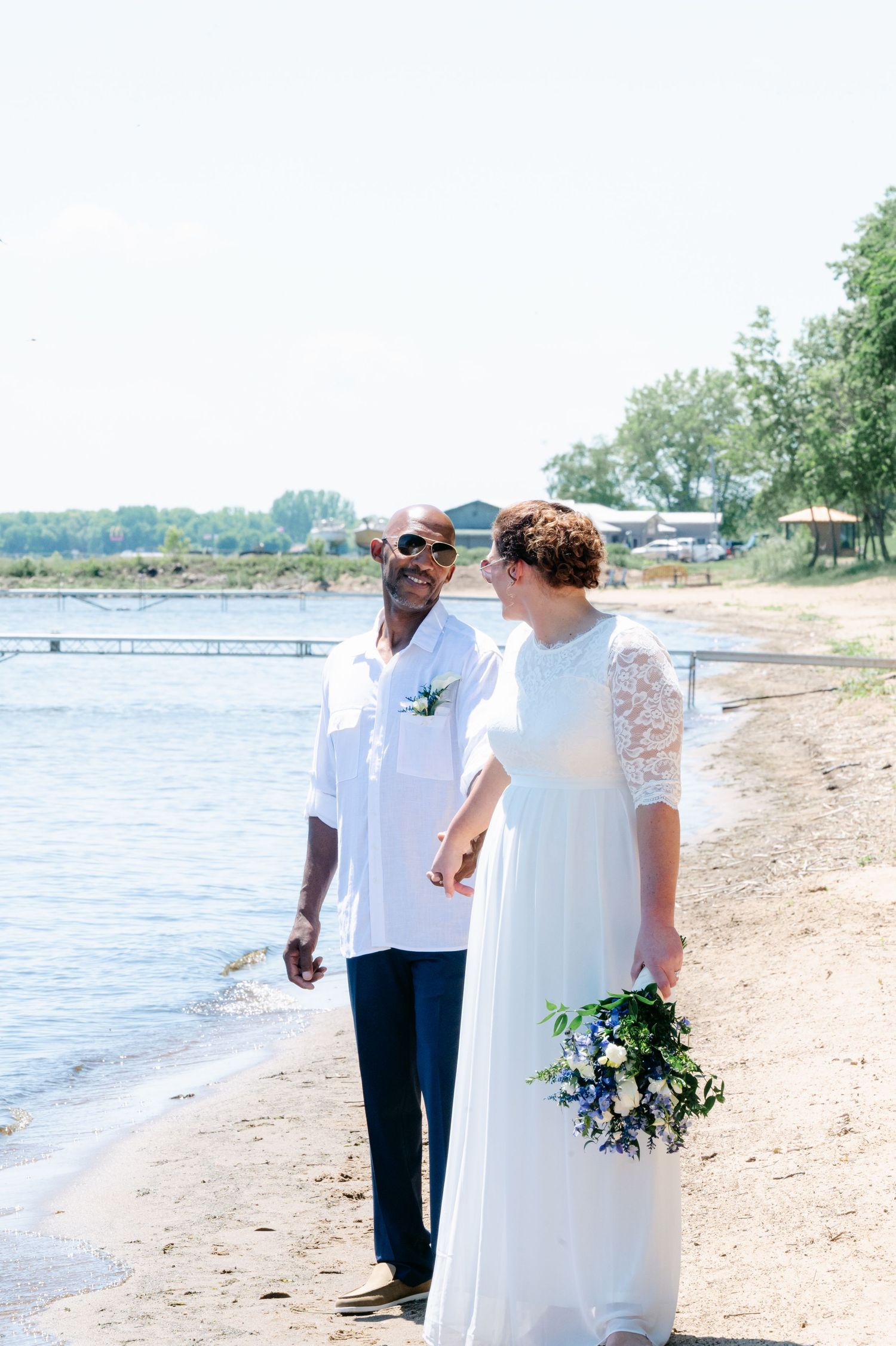 Newlywed interracial couple walking hand in hand on sunny beach in wedding attire.