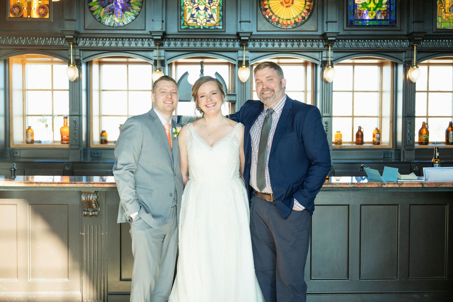 Bride poses with two men in suits inside a vintage bar venue with stained glass windows behind them.
