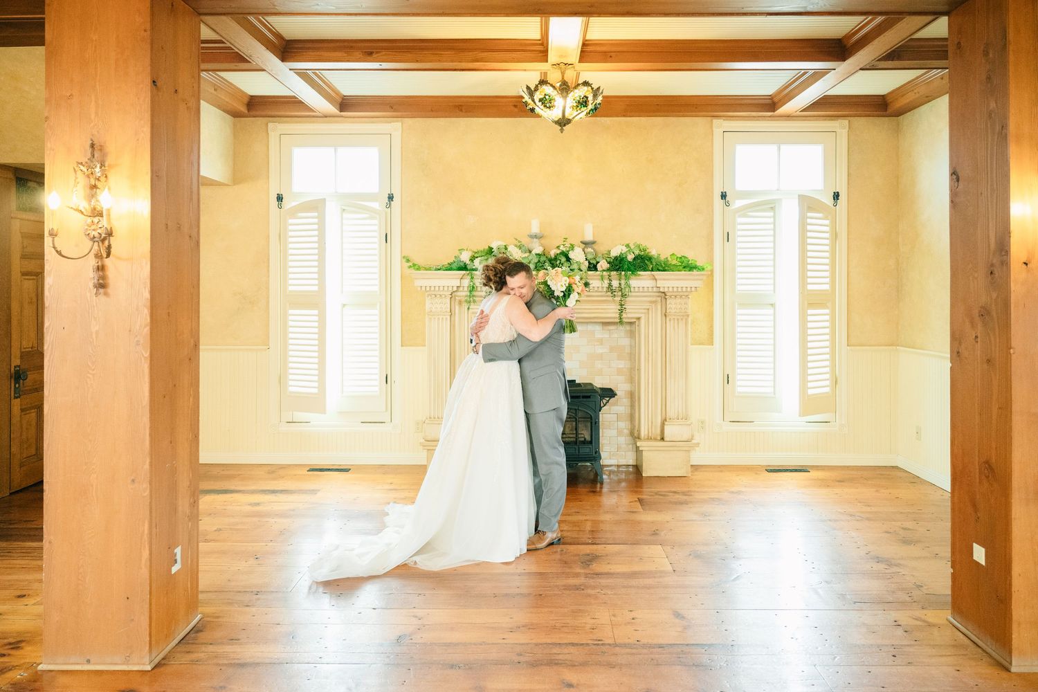 Bride and groom share first dance in elegant wood-floored venue with fireplace and chandelier.
