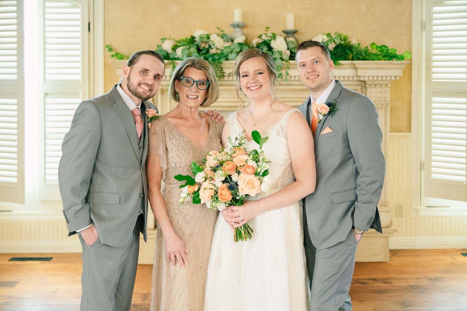 Wedding bride with bouquet poses with family in gray suits and gold dress near floral fireplace.