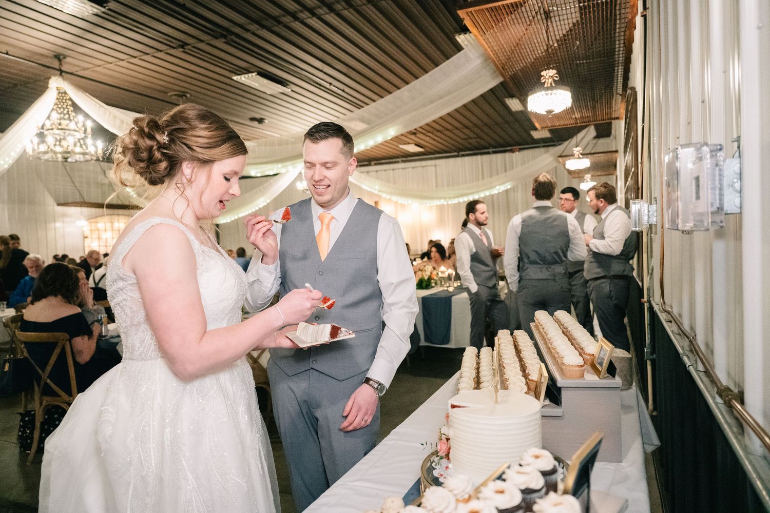 Bride and groom sample wedding desserts at elegant white cake and pastry display table.