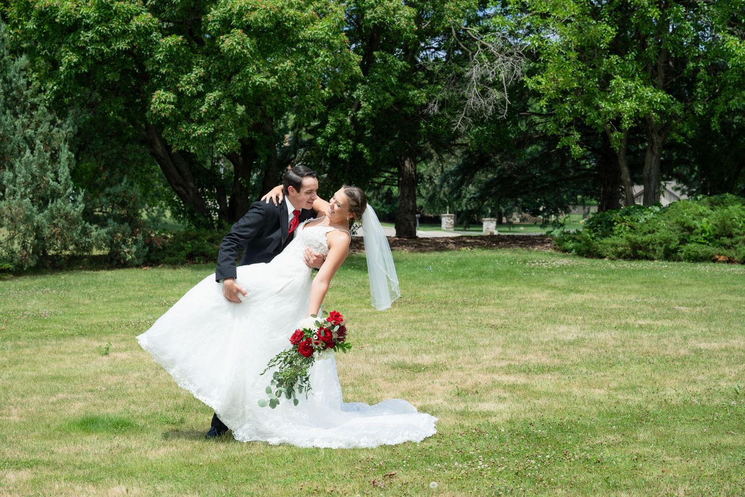 A groom dips his bride for a romantic kiss outdoors on a green lawn with lush trees in the background.