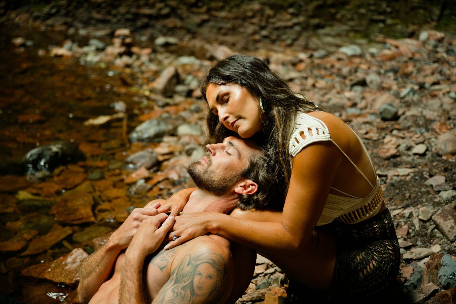 A tattooed shirtless man reclines against rocks outdoors while a woman lovingly holds his face near a stream.