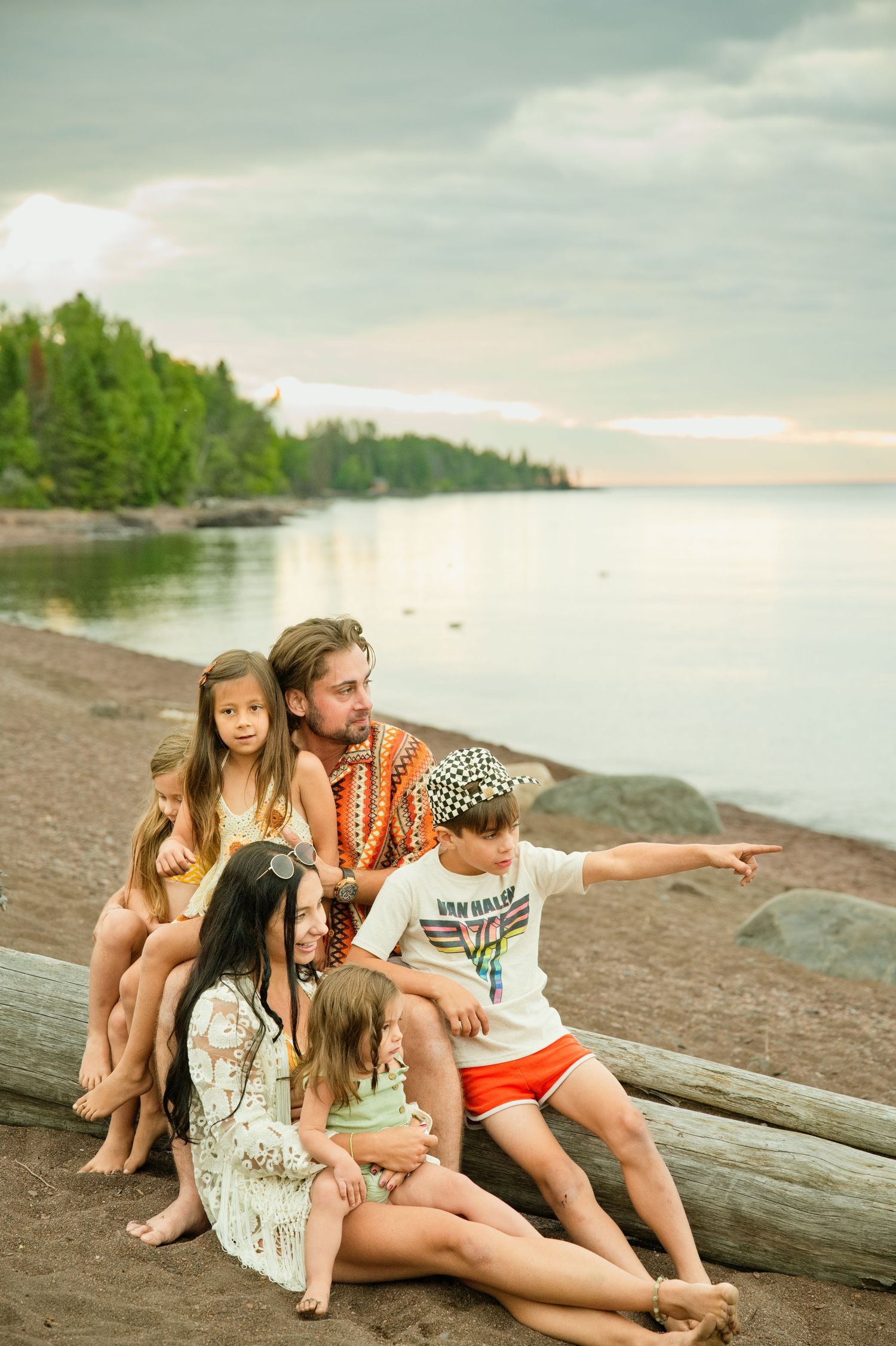Family of five sitting on a driftwood log on a rocky beach at sunset, man pointing toward the horizon.