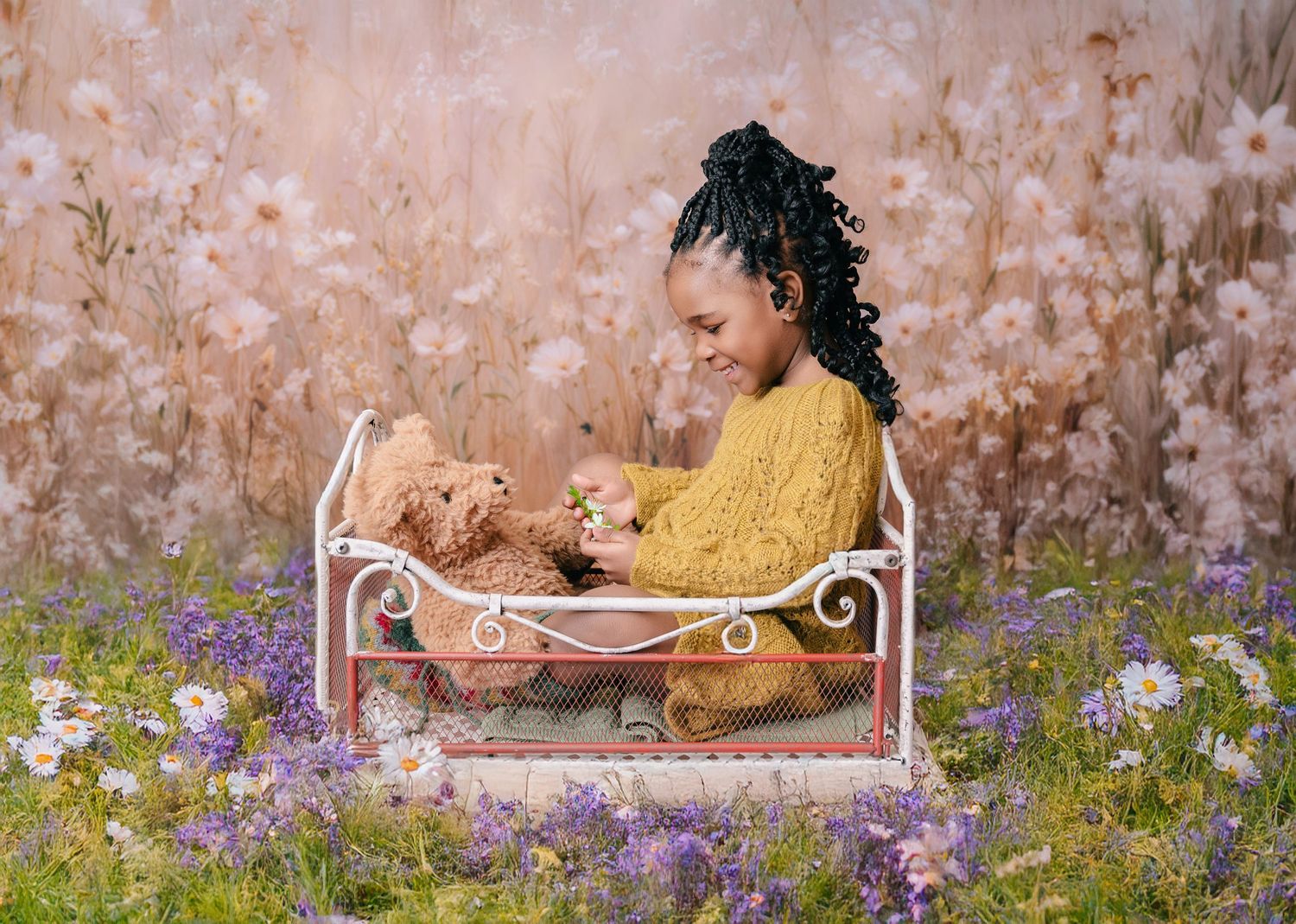 Young girl in yellow knit sweater sits in white mini bed with teddy bear surrounded by purple wildflowers. mother day photoshoot