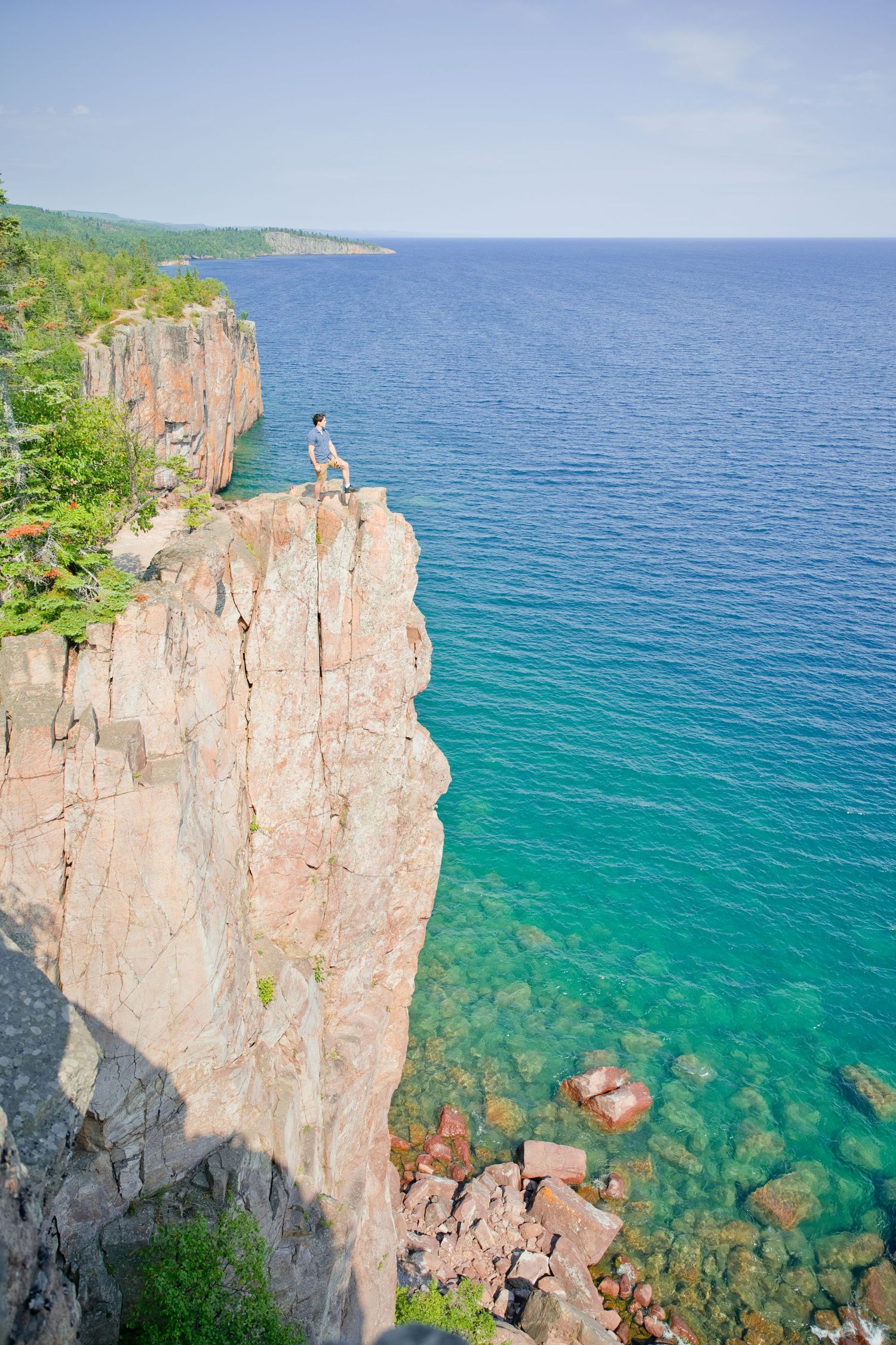 Person standing on edge of dramatic red rock cliff overlooking crystal clear turquoise waters of a vast lake.