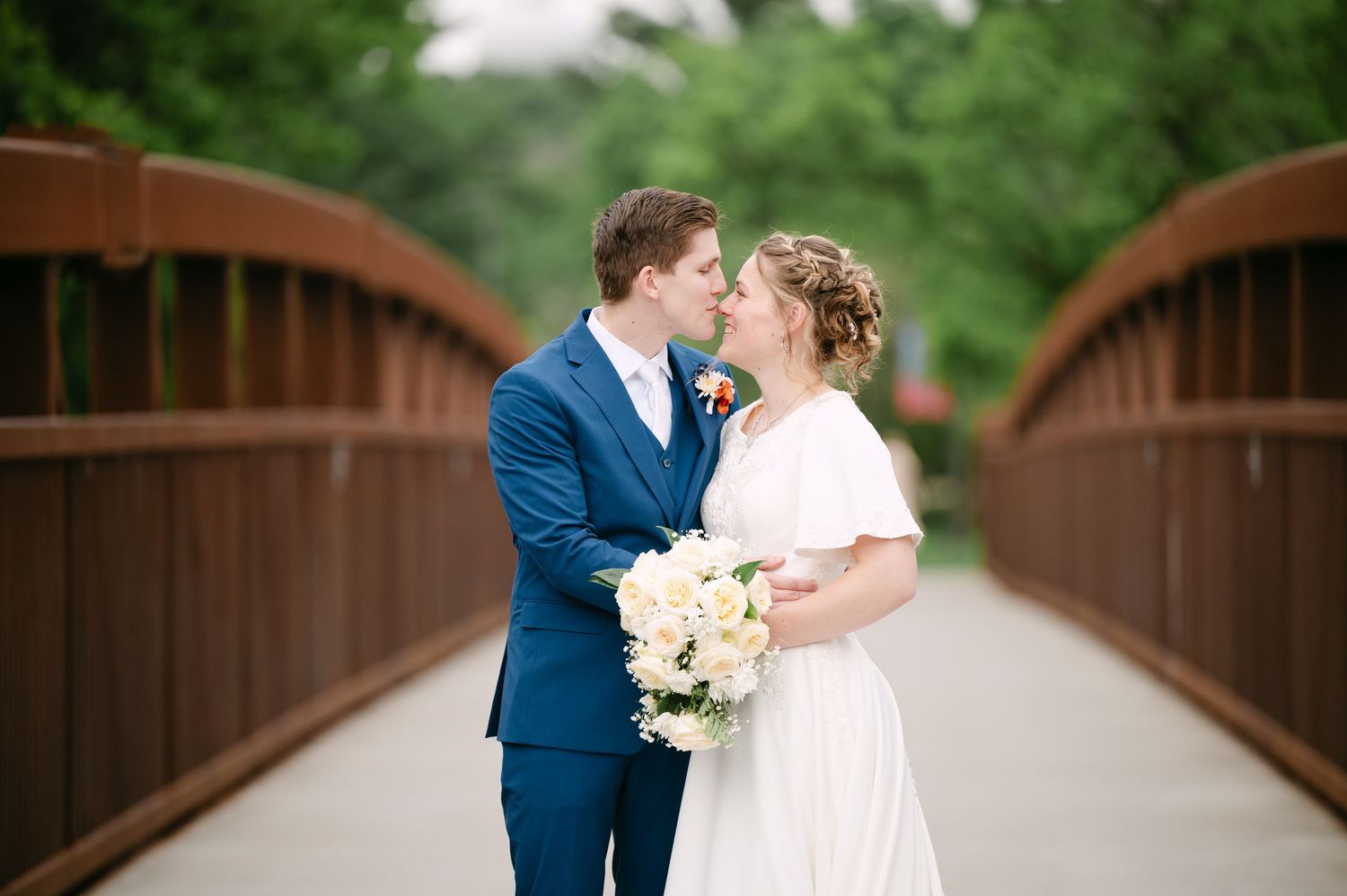 Newlyweds embrace on rustic bridge with white bouquet and navy suits.