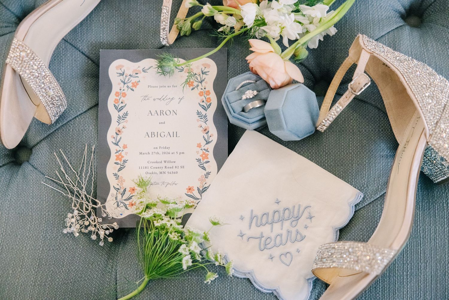 Flat lay of wedding details including floral invitation, glitter heels, happy tears handkerchief, and ring box.