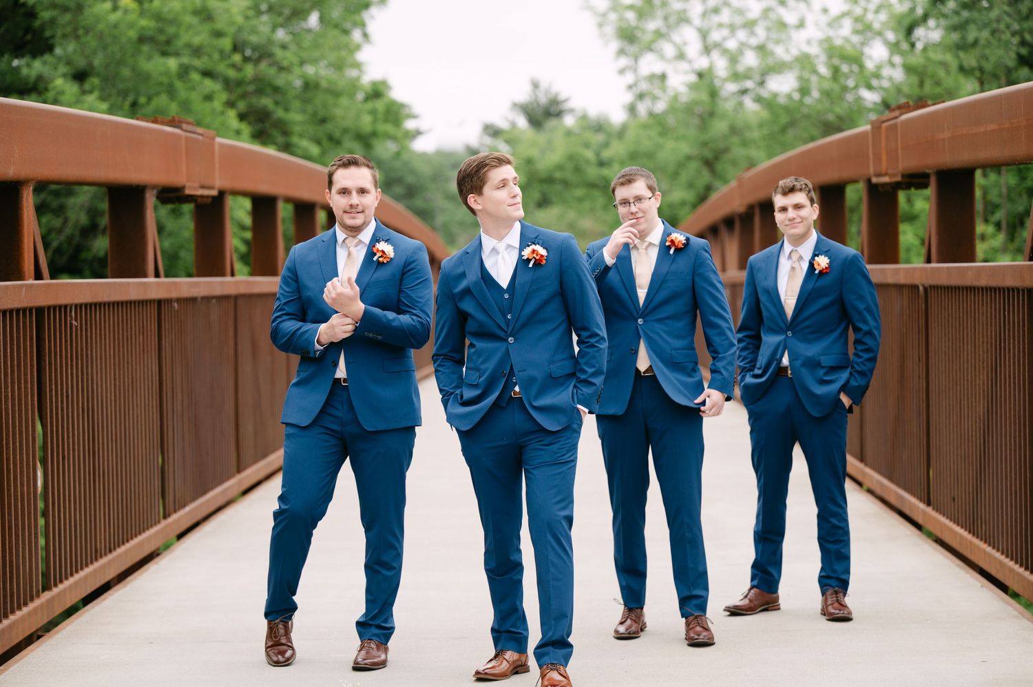 Groomsmen in navy blue suits walking together on a wooden bridge surrounded by greenery.