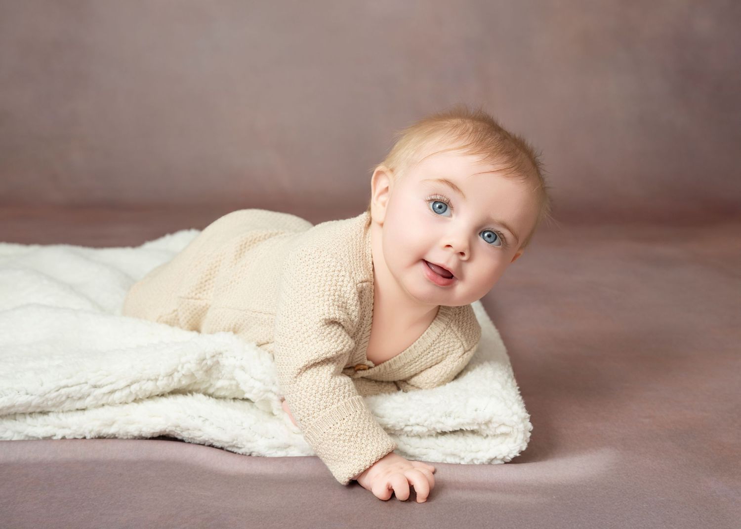 Central MN Milestone Photos - baby with blue eyes lying on a white blanket, wearing a cream knit outfit against a soft pink background.