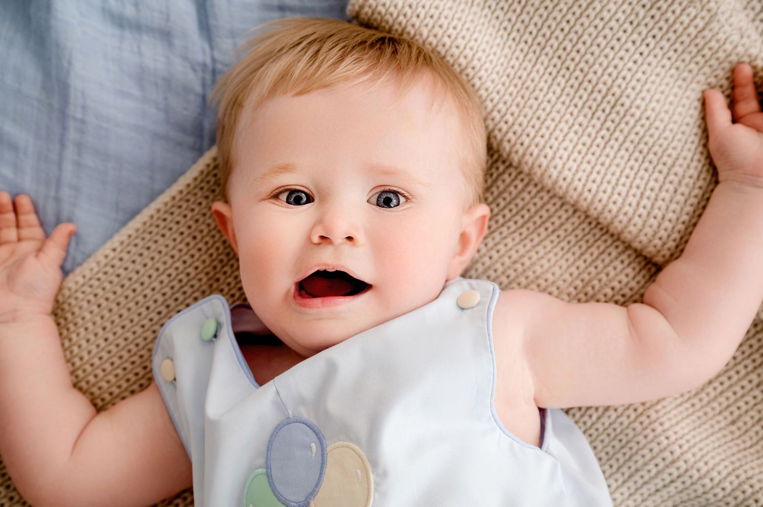 Central MN Milestone Photography - Adorable baby lying on a knit blanket wearing a white sleeveless outfit, looking up with mouth open.