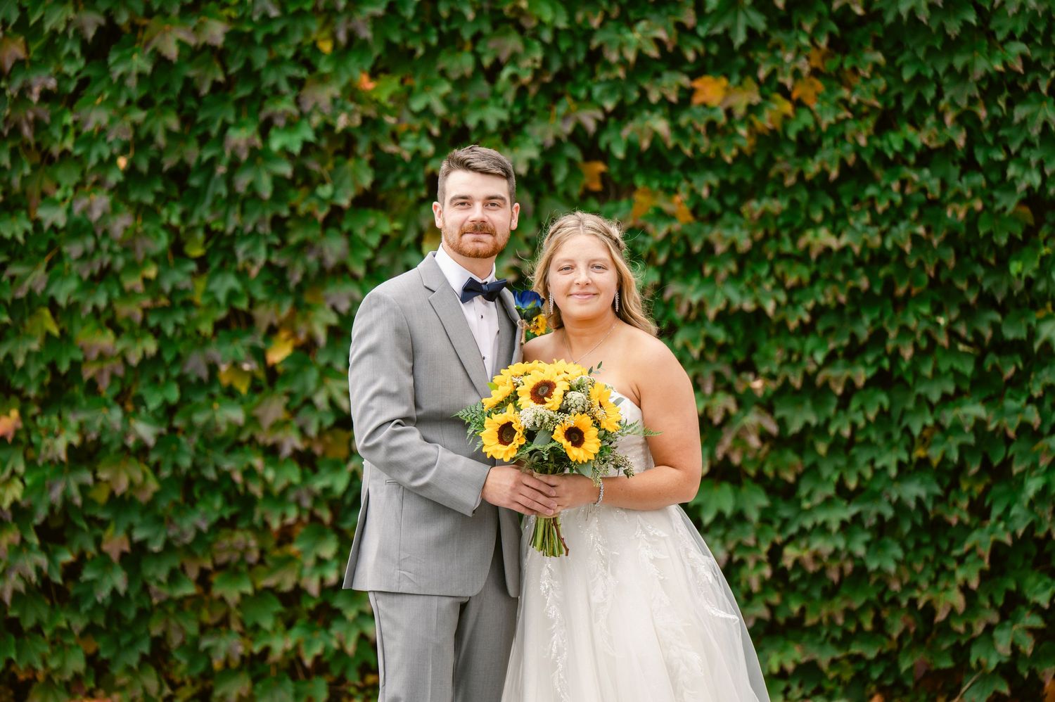 Central MN Wedding Photos | Bride and groom posing together in front of a lush green ivy wall with sunflower bouquet.