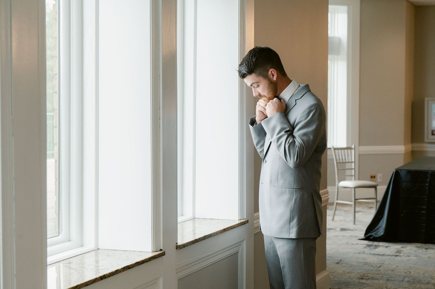 Central MN Wedding Photos | Groom in grey suit standing by large windows, adjusting his cufflinks before the wedding ceremony.