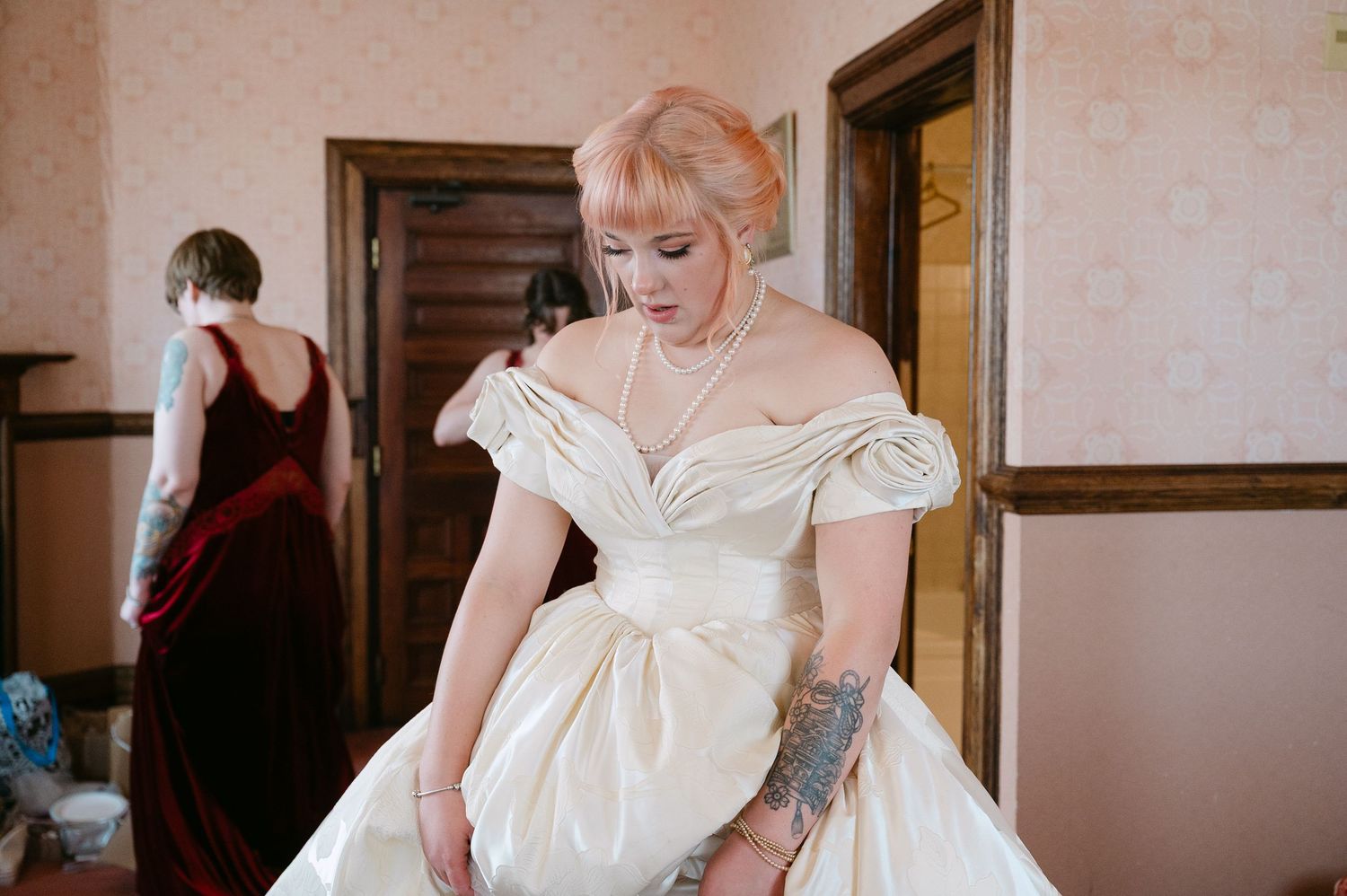 Central MN Wedding Photos | Bride in elegant off-shoulder ivory wedding gown with tattoos adjusting dress in vintage interior.
