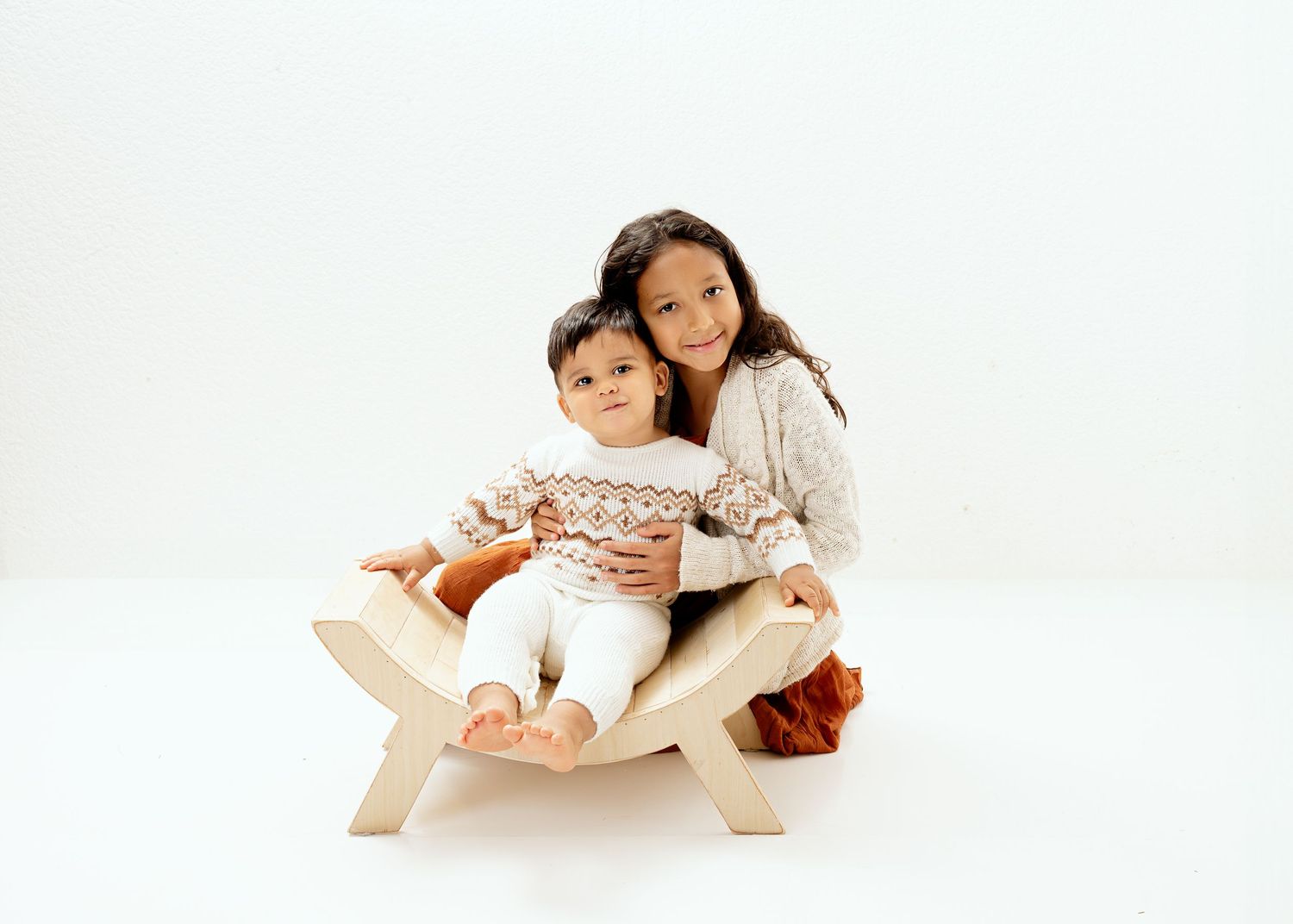 Two children sit together on a wooden bench against a white studio background.
