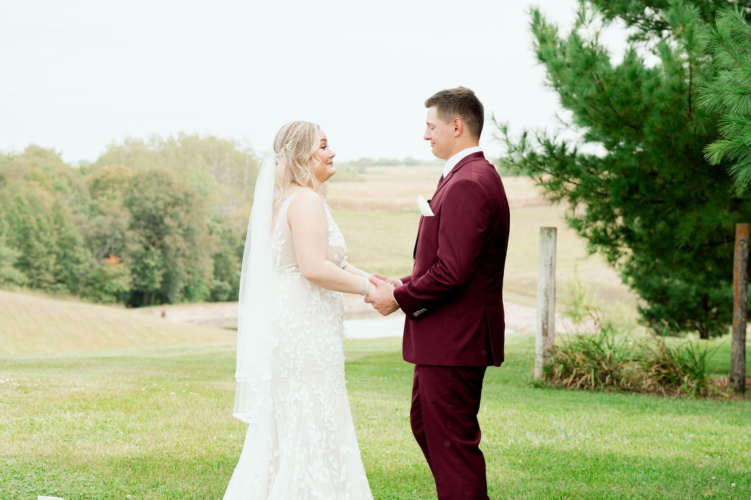 A bride and groom share a tender moment in a rustic outdoor setting with green trees in the background.