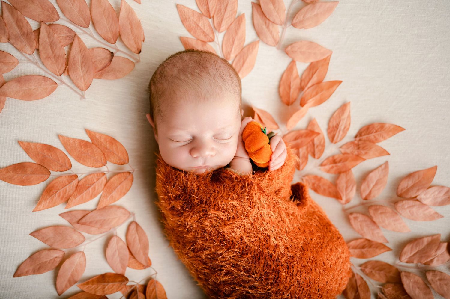 Sleeping newborn wrapped in soft orange fabric surrounded by delicate peach-colored leaf decorations against white background.