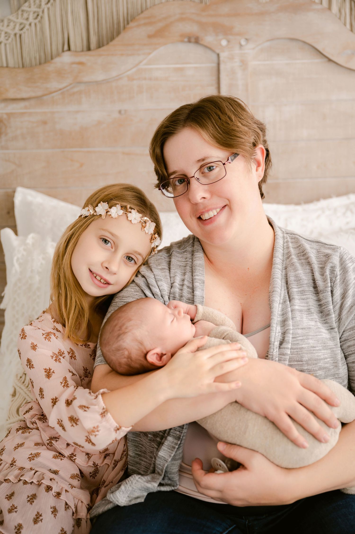 A family portrait showing a mother in gray holding a newborn baby while sitting with a young girl wearing a floral crown.