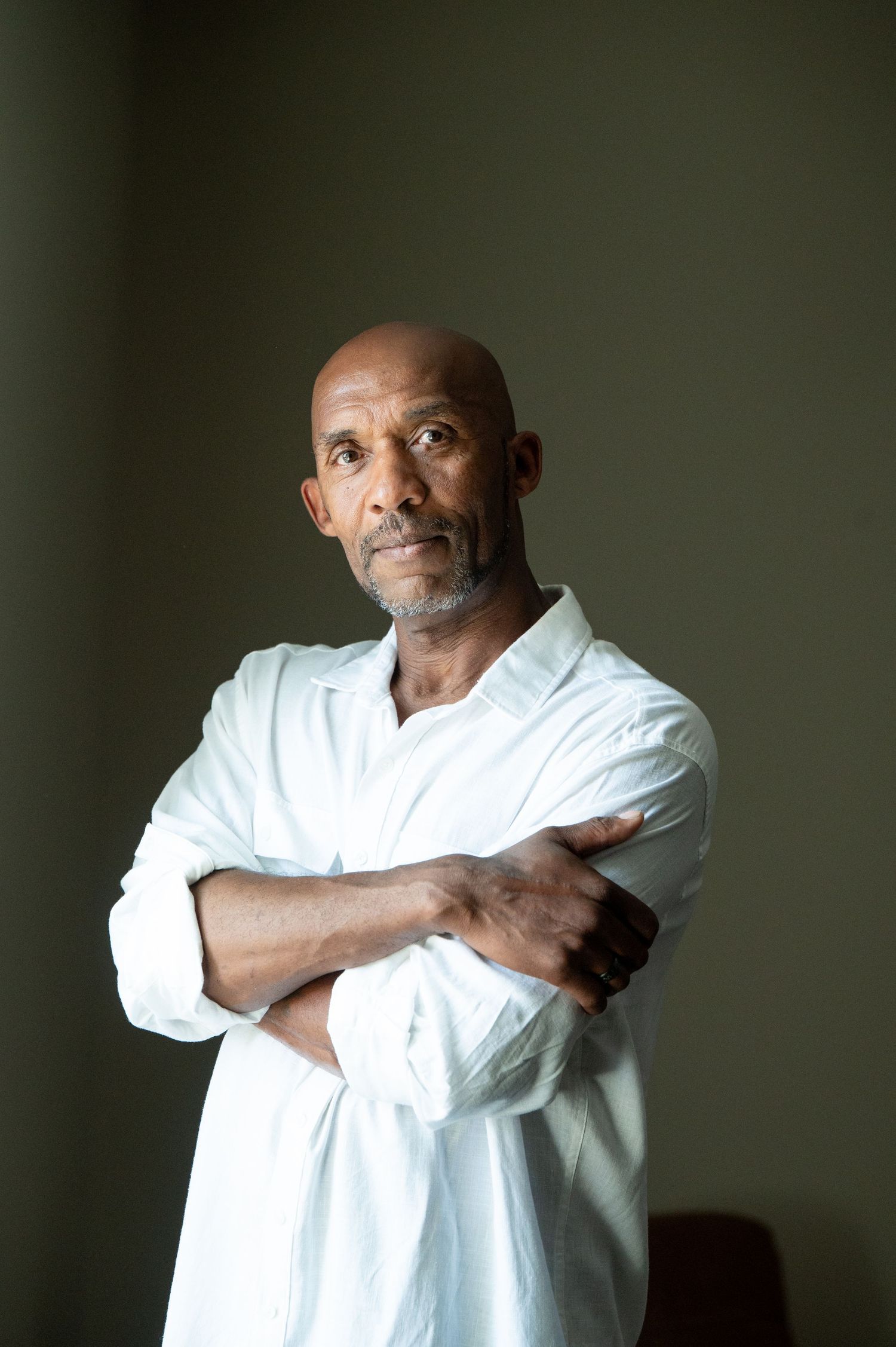 Bald man with gray beard in white shirt stands with arms crossed against dark background.