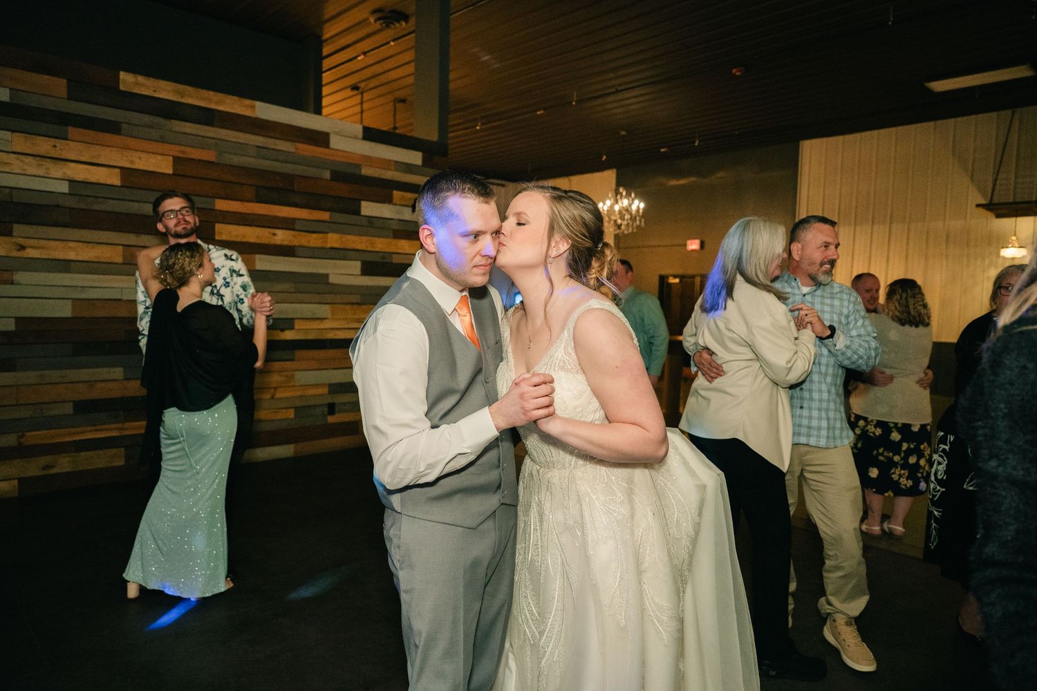 Bride and groom share a kiss on the dance floor surrounded by wedding reception guests.