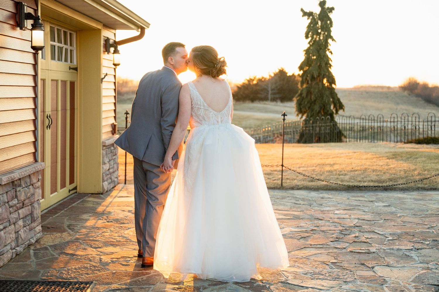 Bride and groom share a sunset kiss holding hands outside a rustic wedding venue.
