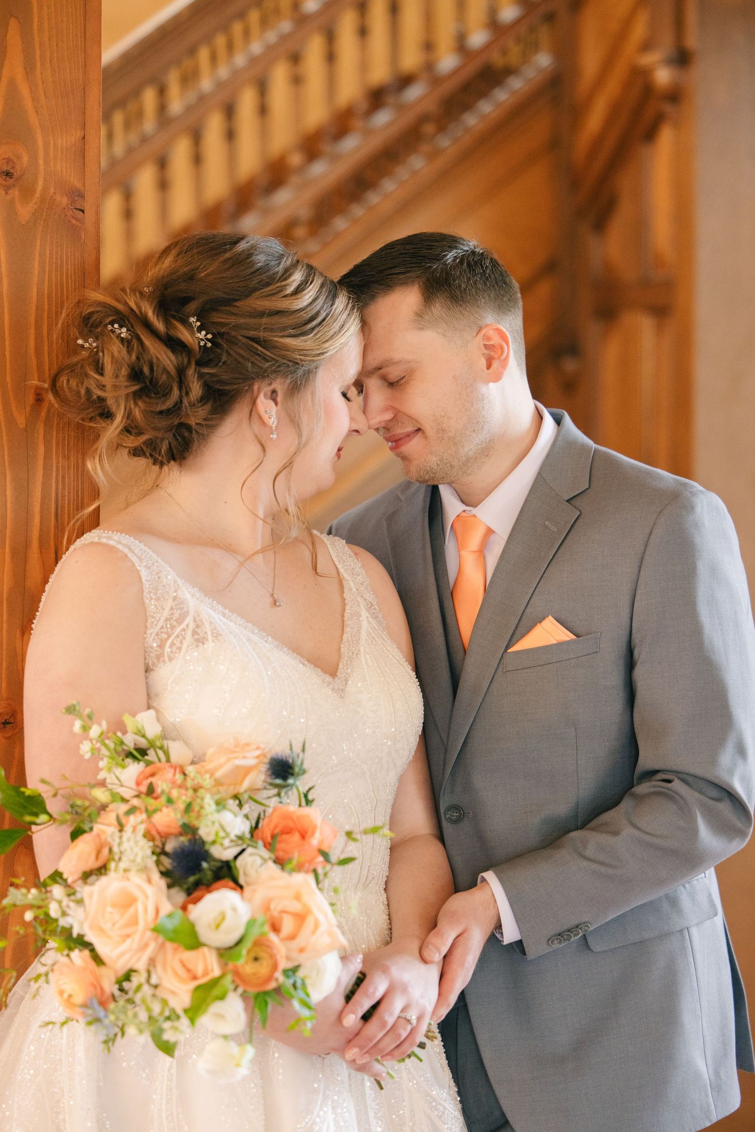 Bride and groom share a romantic forehead touch, bride holding a peach floral bouquet in a wooden venue.