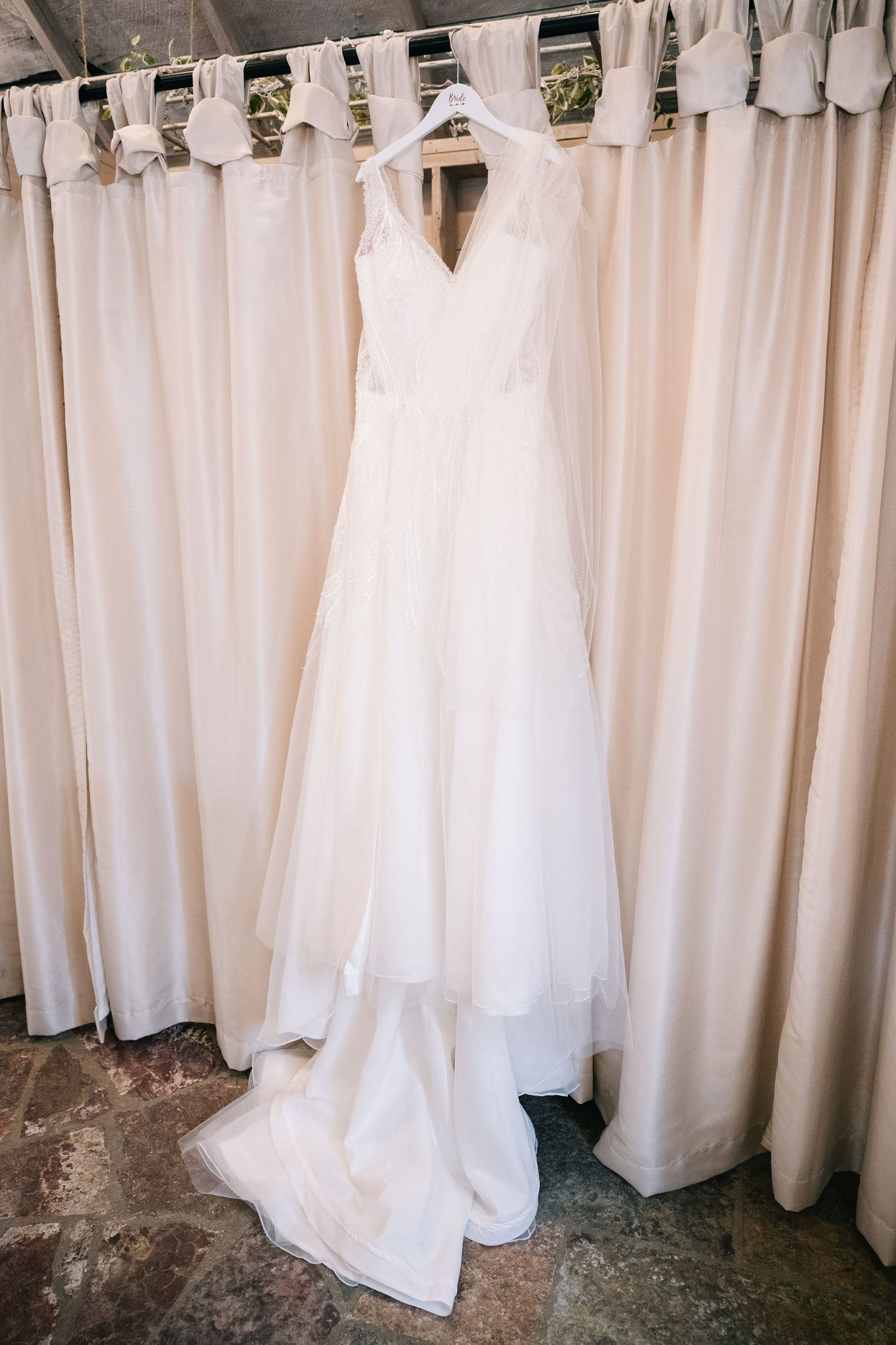 Elegant white A-line wedding dress with lace details hanging on a hanger against cream curtains.
