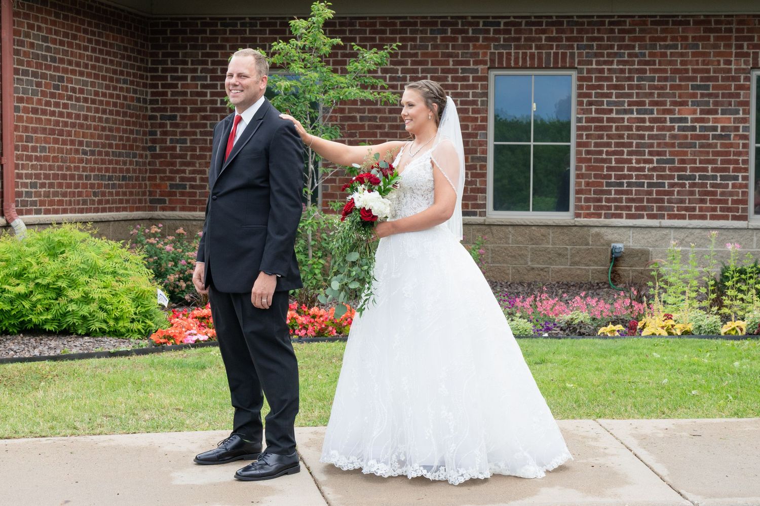 Bride in white lace gown holding red bouquet stands beside groom in black suit outside brick building.