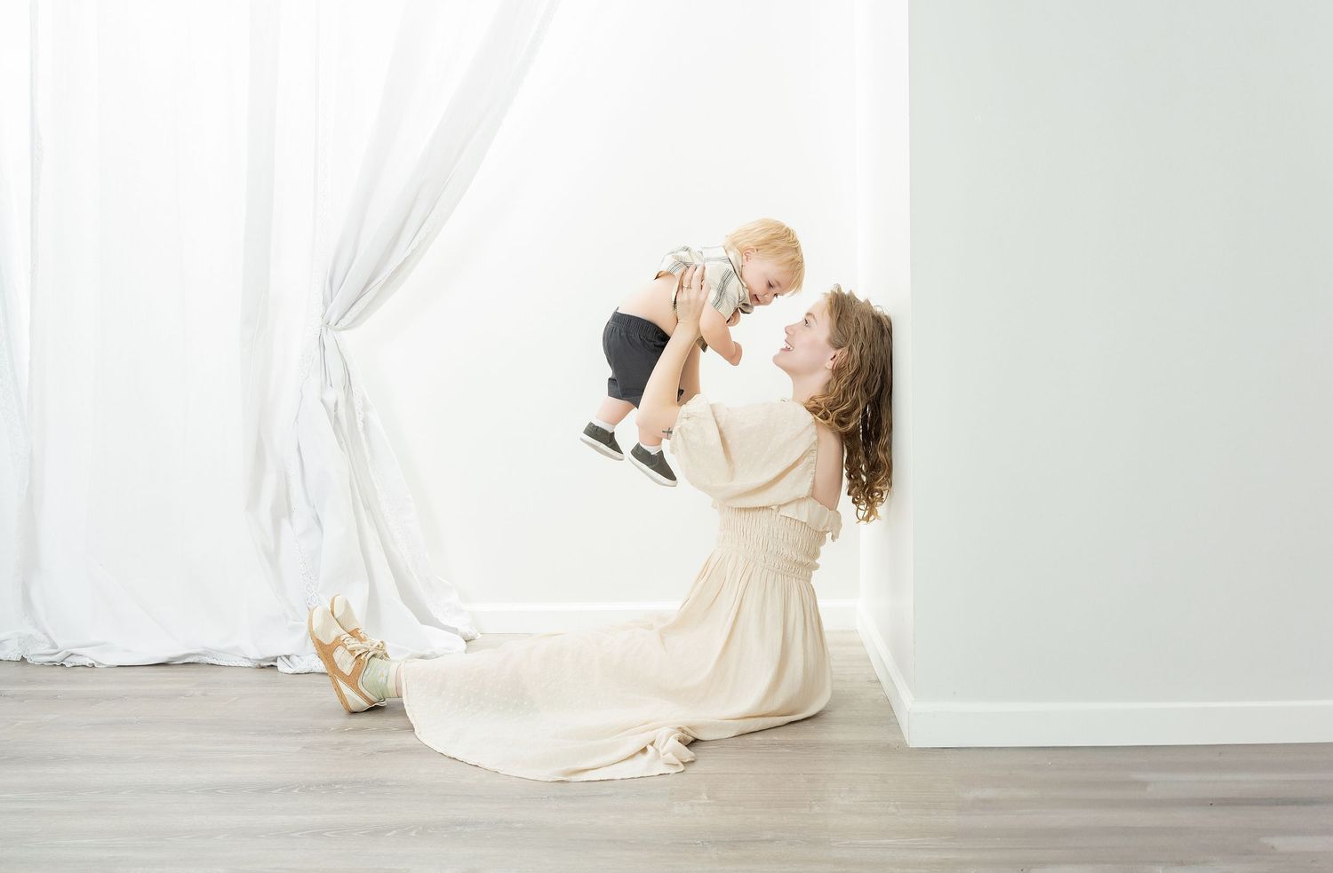 A ballerina in a flowing cream dress holds a baby while dancing in a bright minimalist studio setting. Princeton MN Milestone, Maternity and