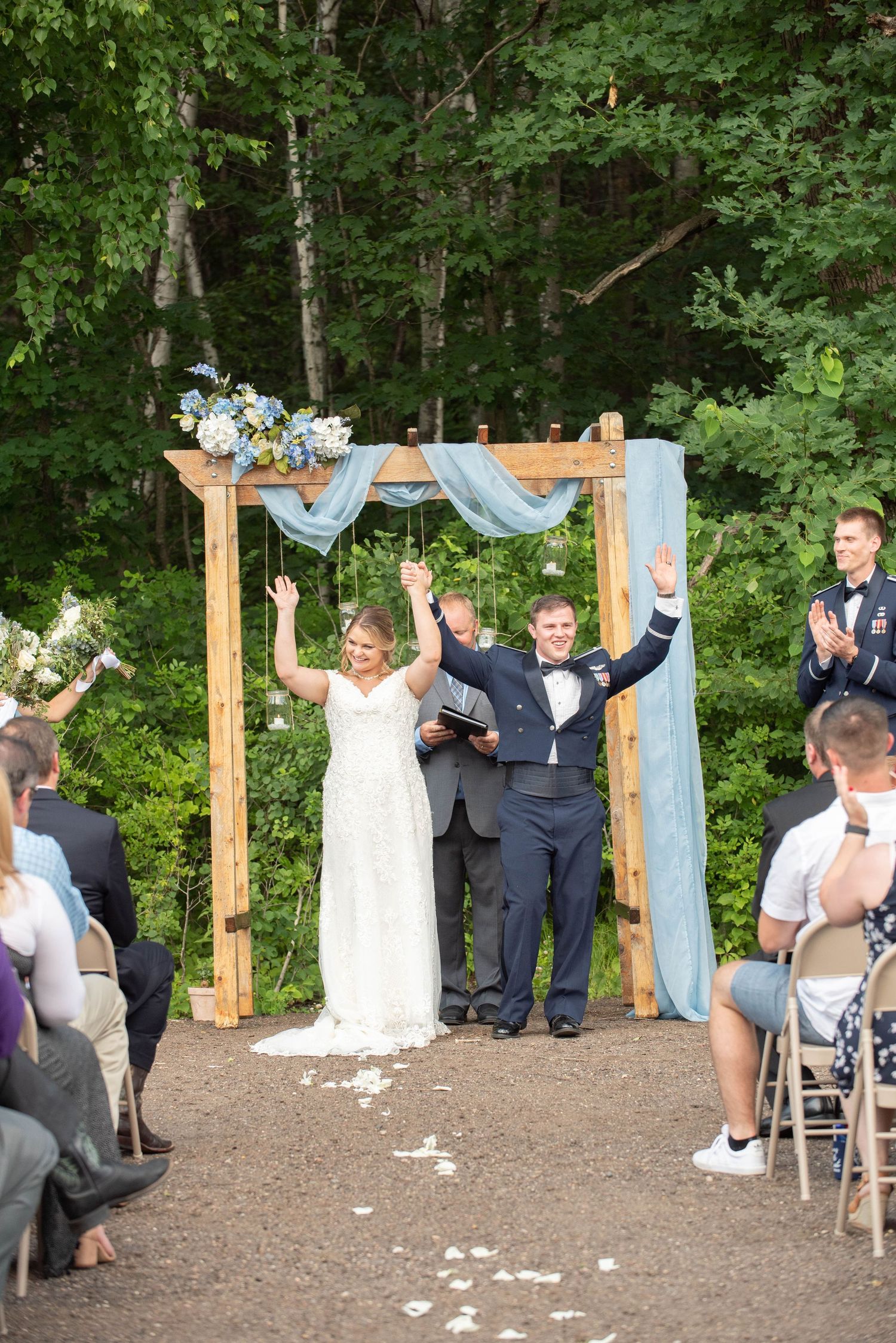 Celebratory moment under wooden wedding arch as guests seated on folding chairs applaud. Zimmerman MN Weddings
