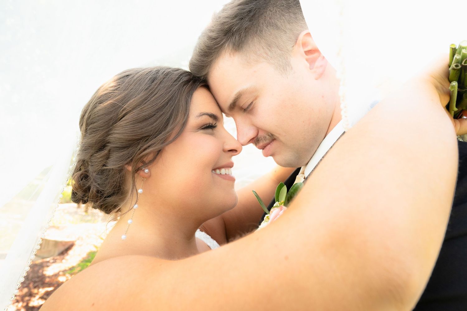 Bride and groom share a romantic forehead-to-forehead moment wrapped in a white veil outdoors.