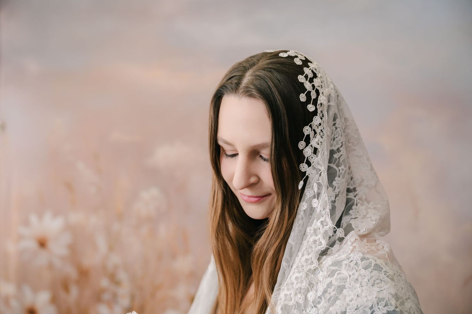 Young woman in white lace veil and floral crown gazes down softly in a dreamy outdoor portrait. mother day photoshoot